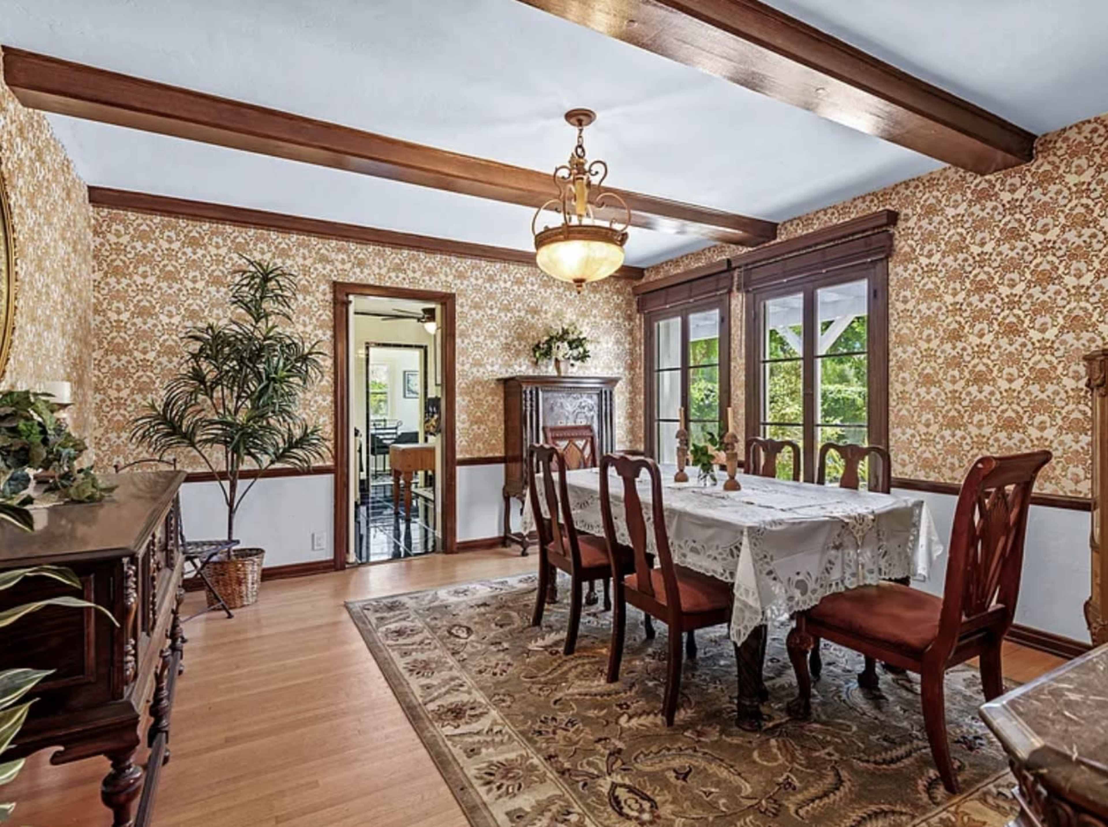 The image shows a dining room featuring a large wooden table surrounded by eight chairs, with floral wallpaper and wooden beams on the ceiling.