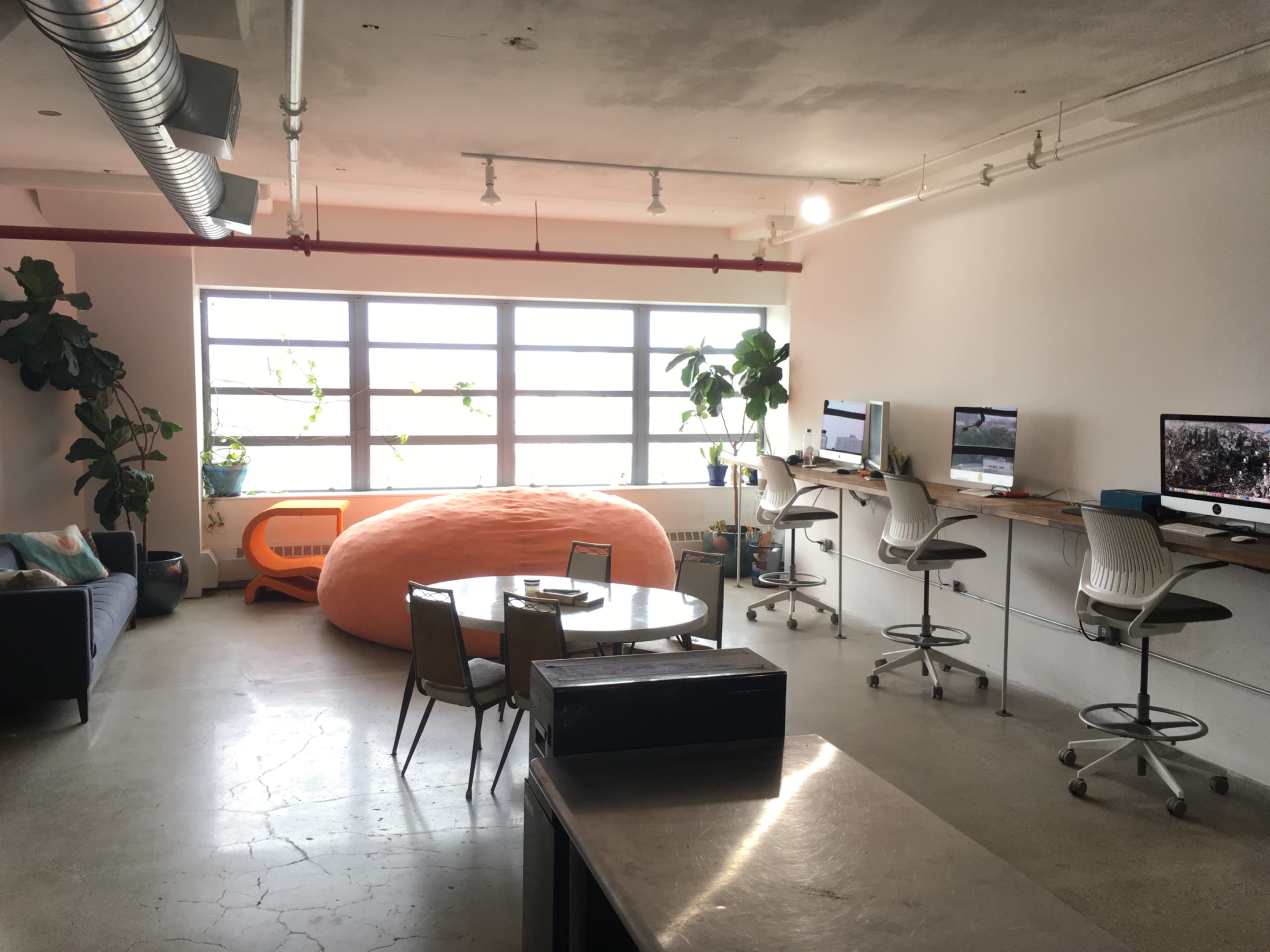 The image shows a modern, open office space featuring a large orange bean bag, a round dining table, and several workstations with computers, all illuminated by natural light from large windows.