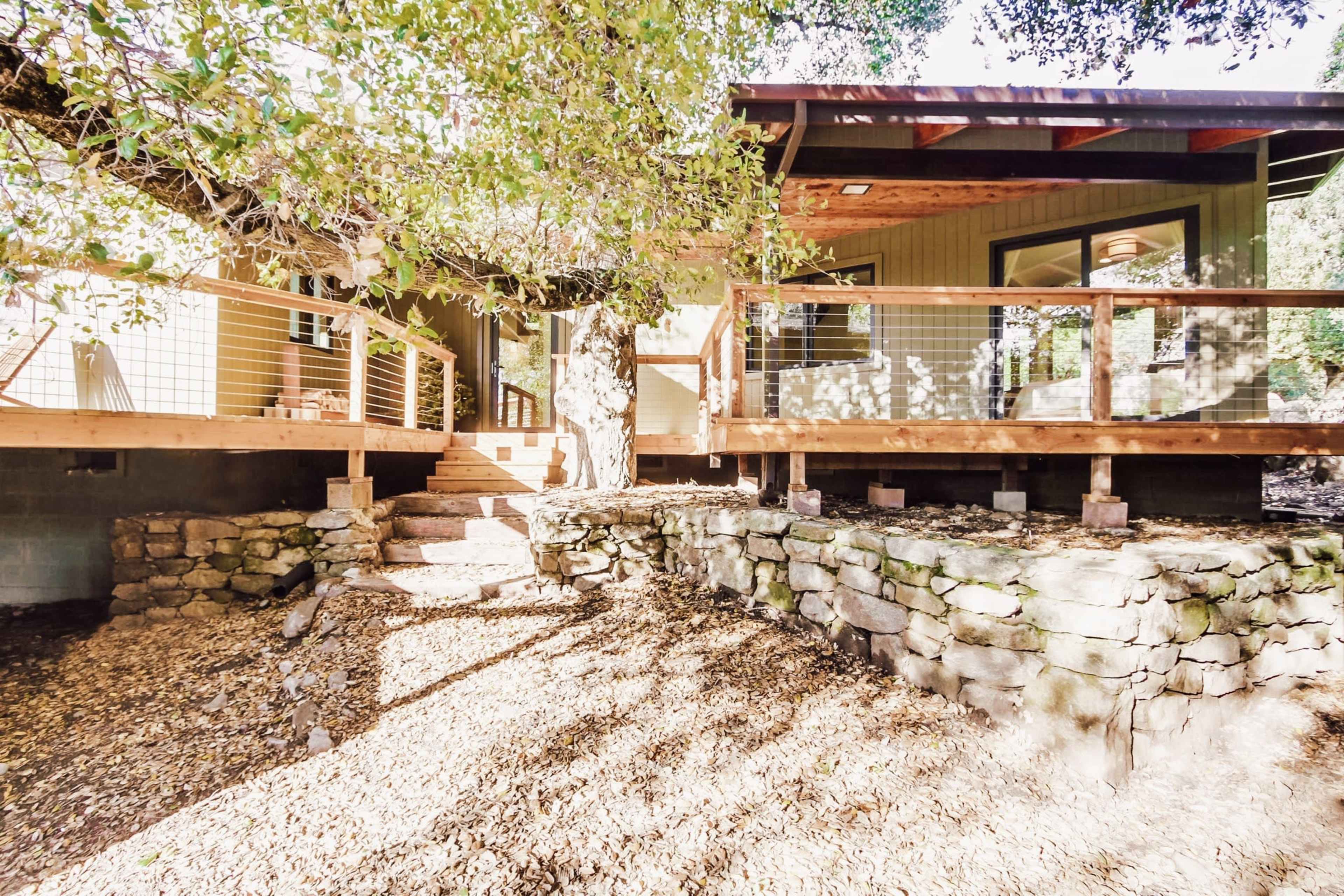 A house elevated on stone supports with wooden decks and steps leading up, surrounded by gravel and trees.