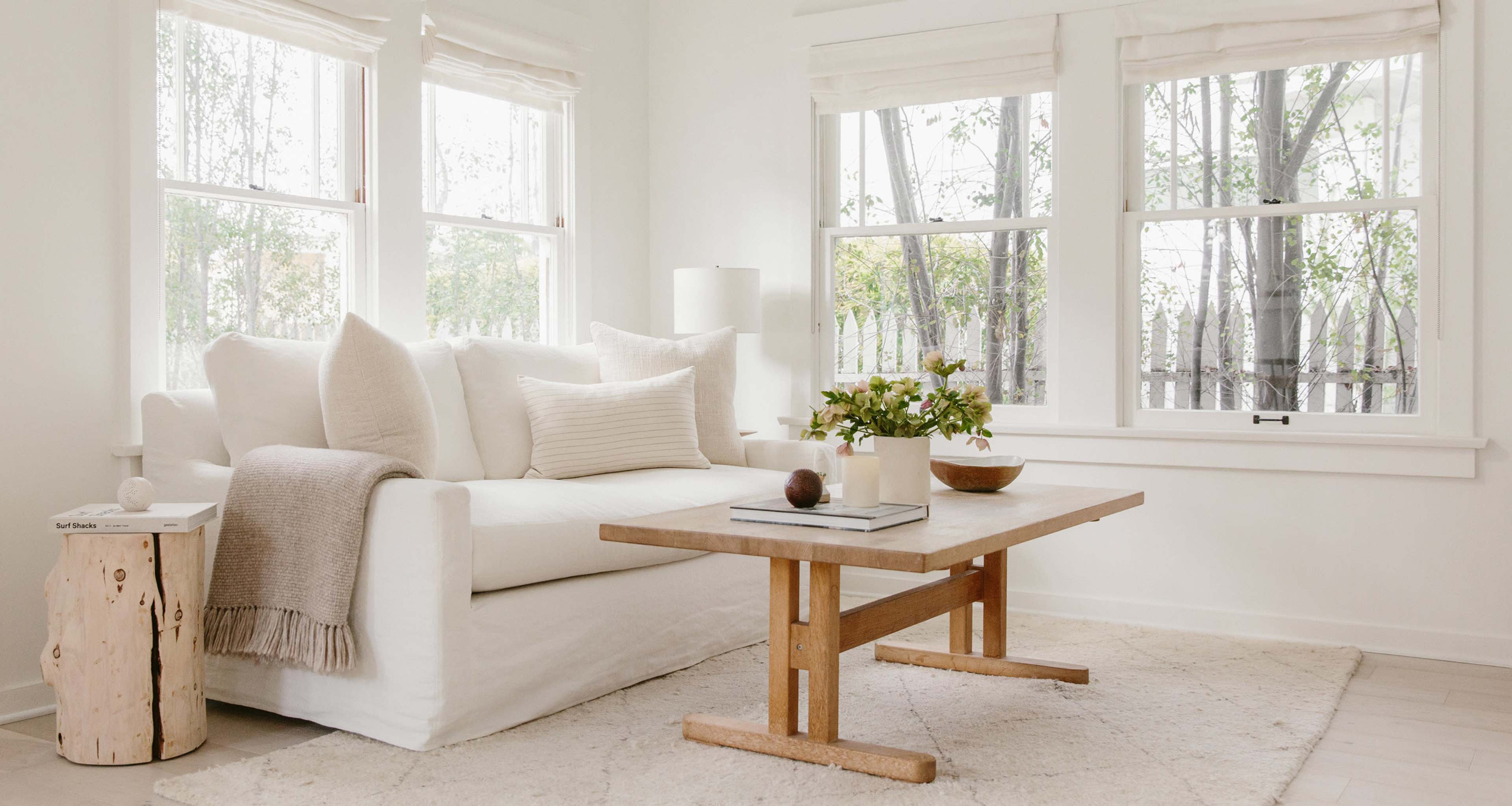 A bright living room features a white couch, a wood coffee table, and large windows letting in natural light.