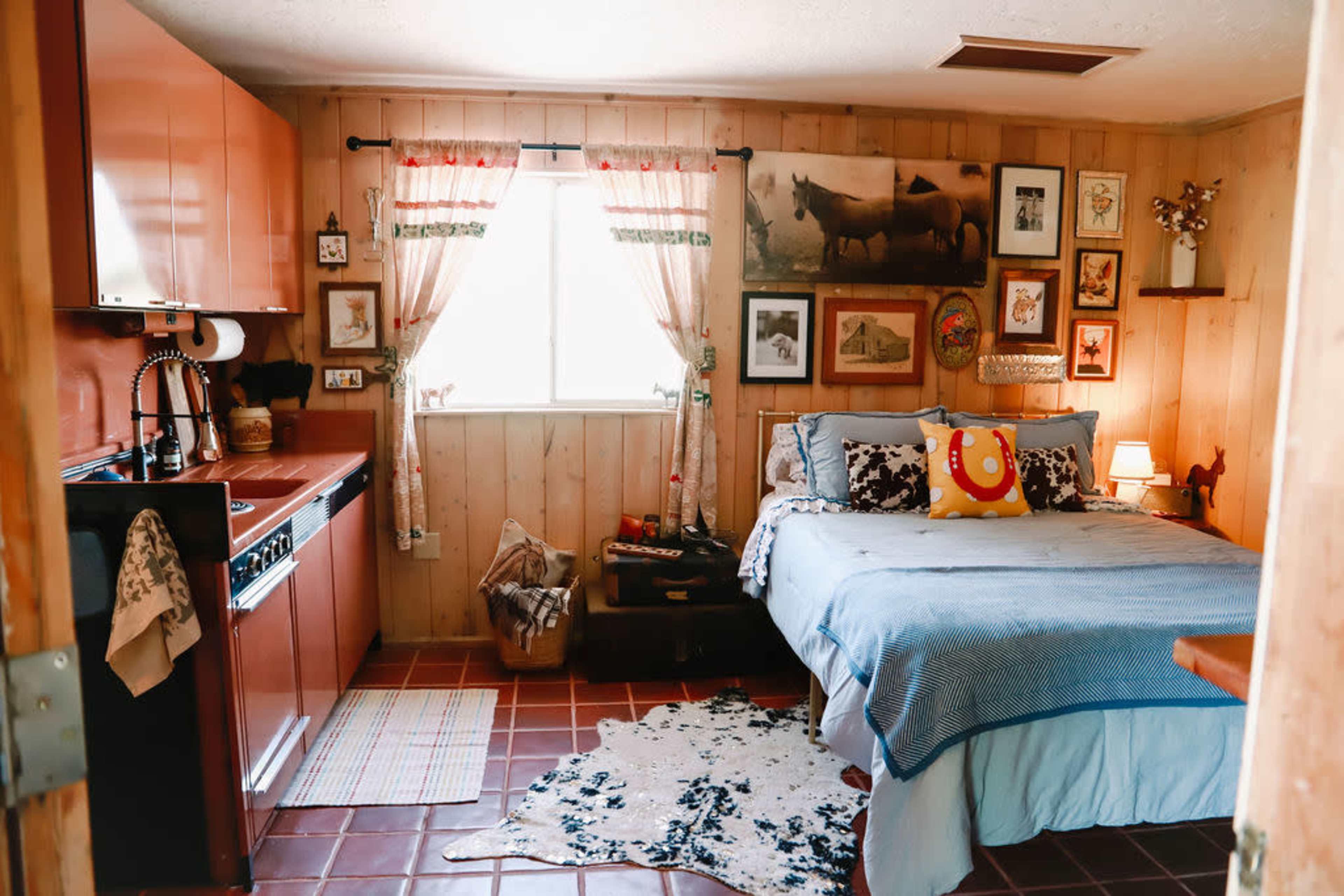 The image shows a cozy bedroom with wooden walls, a bed with blue bedding, and a kitchen area featuring red cabinets.