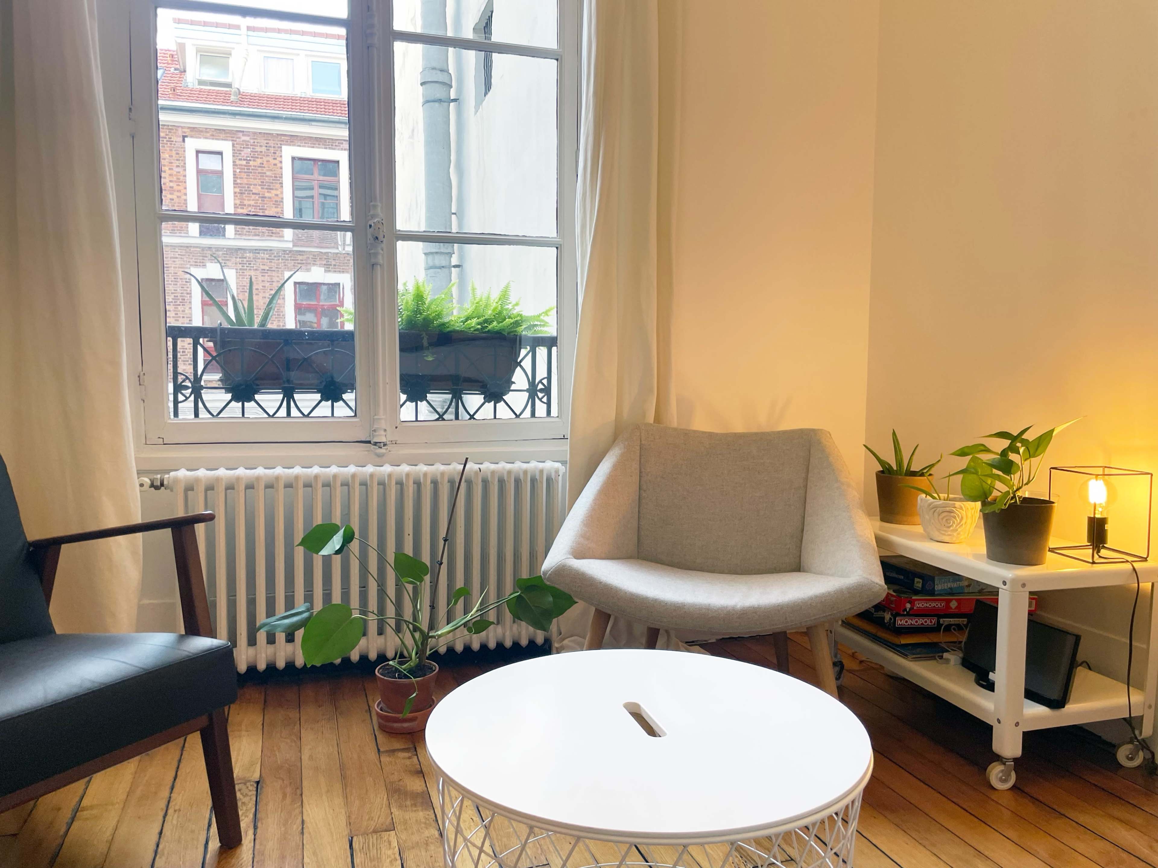 The image shows a cozy room with a black chair, a light gray chair, a round white coffee table, a nearby plant, and a window with potted plants on the sill.