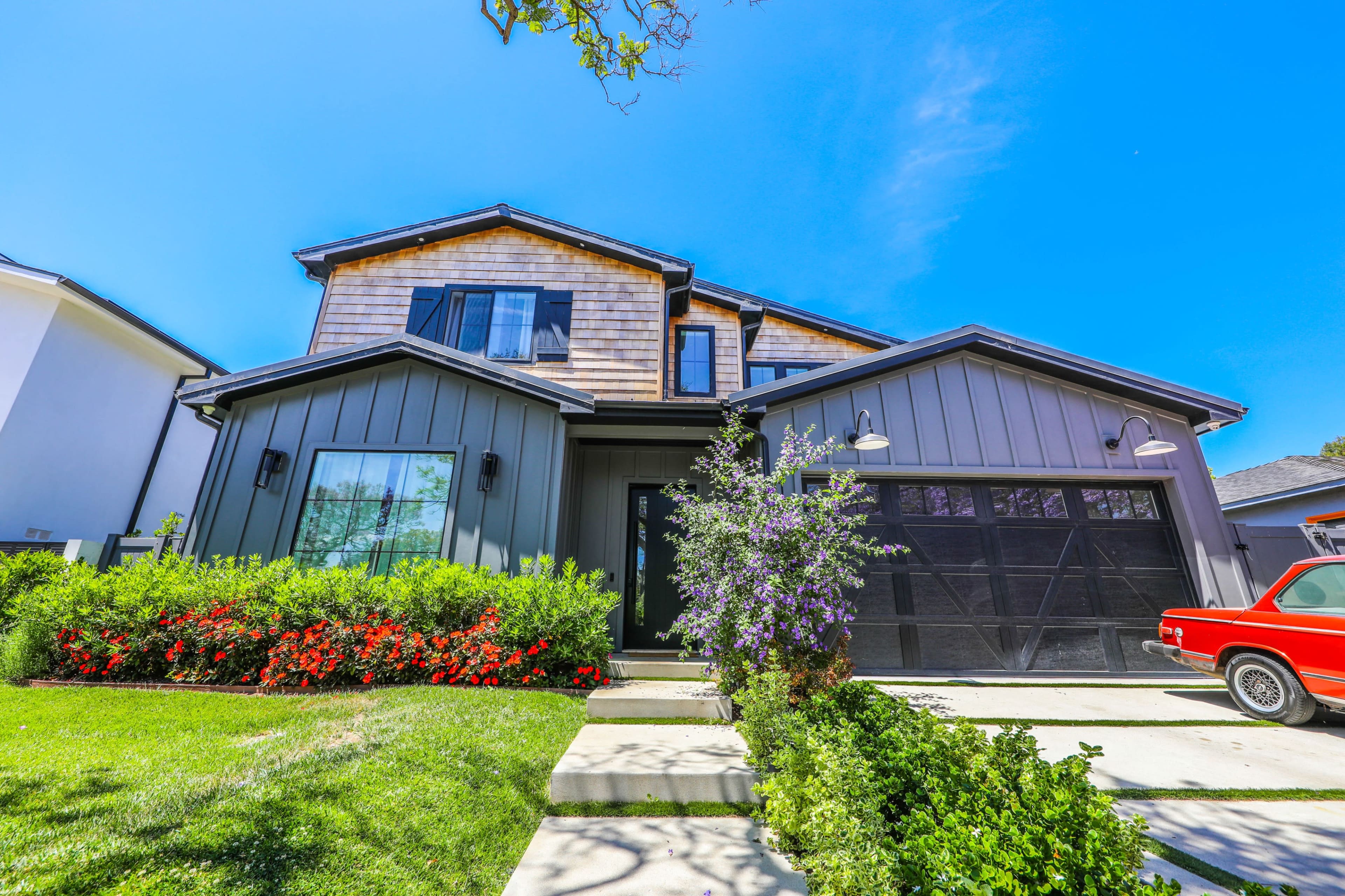 A two-story modern-style house with a gray exterior, a large front yard featuring colorful flowers, and a red car parked in the driveway.