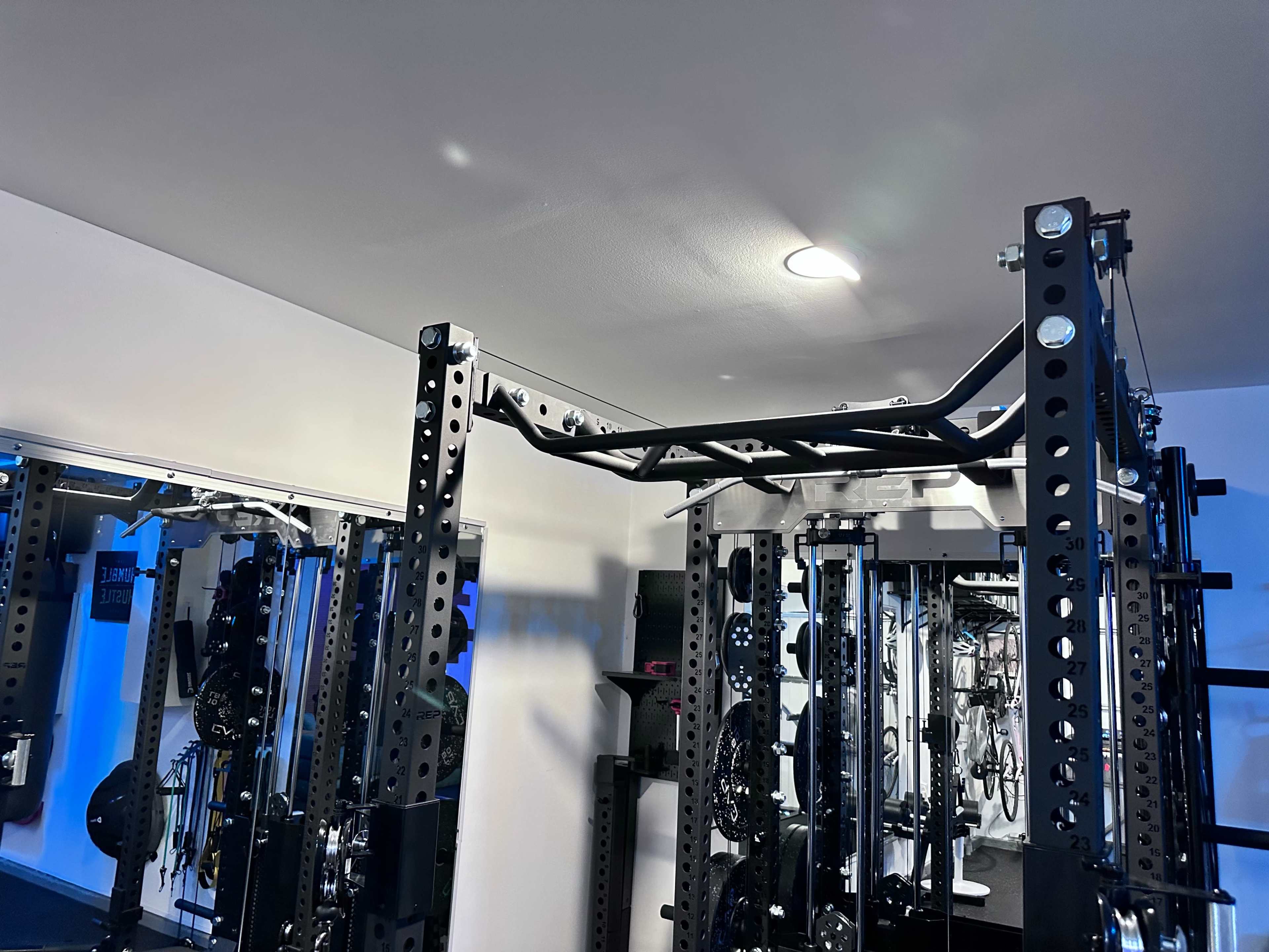 The image shows a corner of a gym featuring a squat rack with a pull-up bar and various gym equipment in the background.