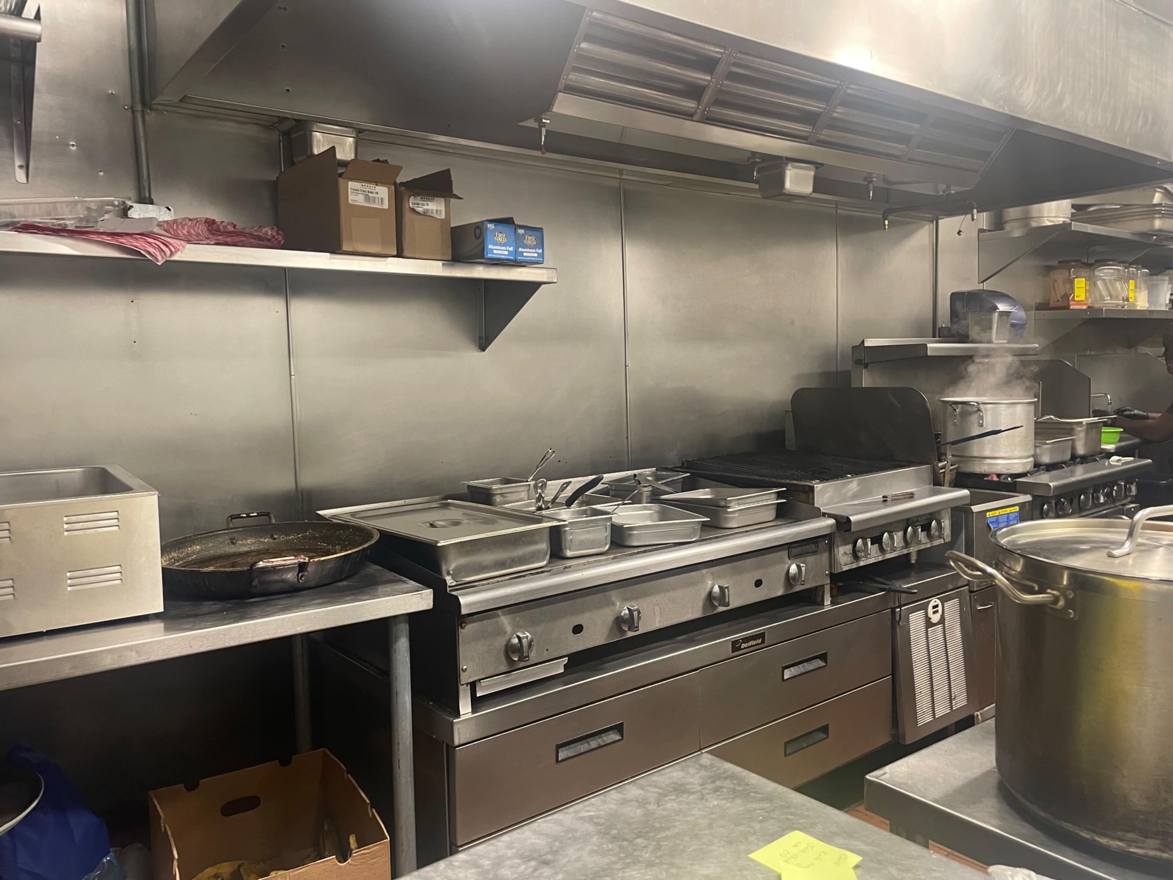 A commercial kitchen with stainless steel equipment including a stove, pots, and utensils on shelves.