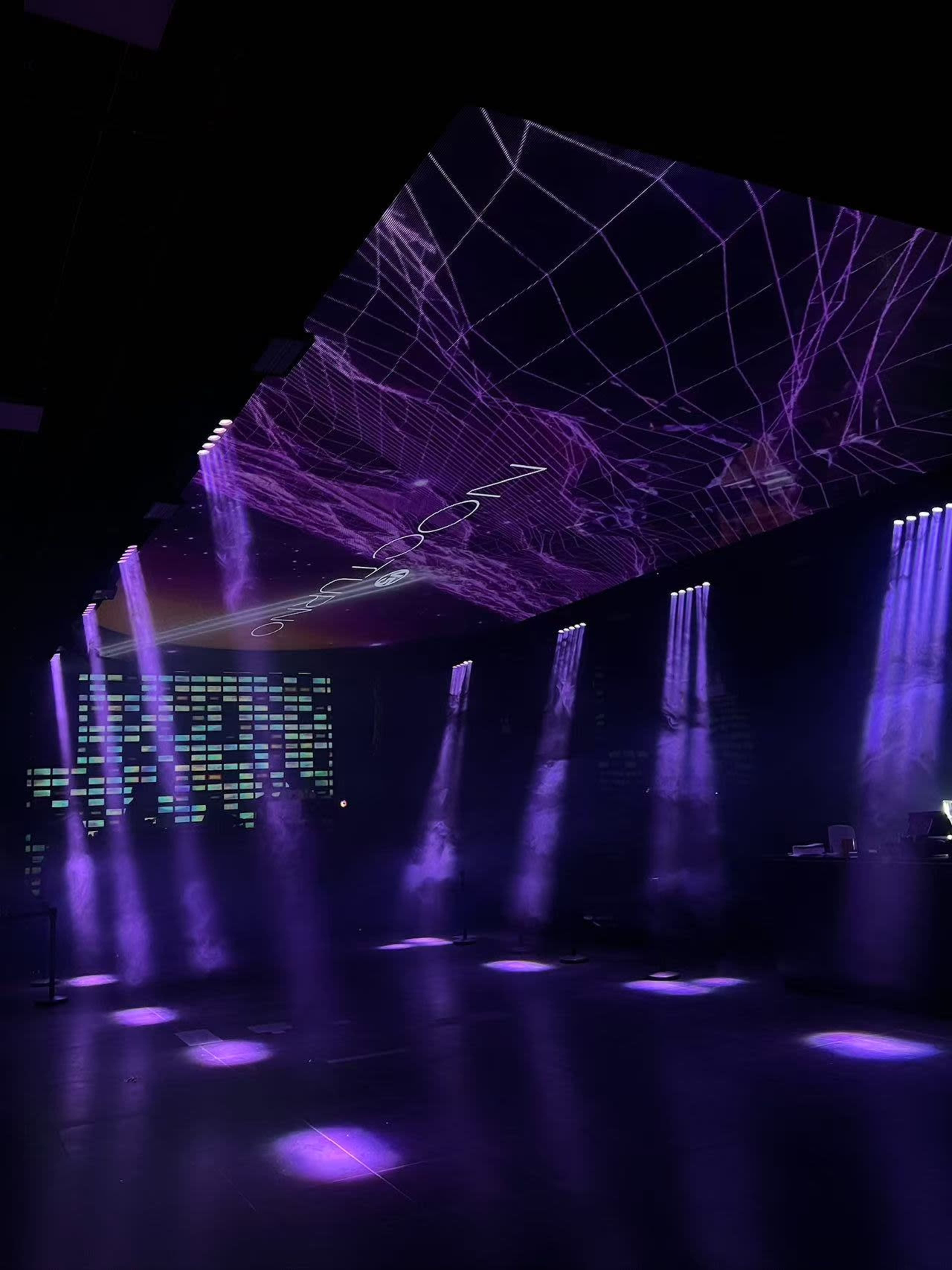 A dark room features purple lighting and projected graphics on the ceiling, with beams of light illuminating the floor.