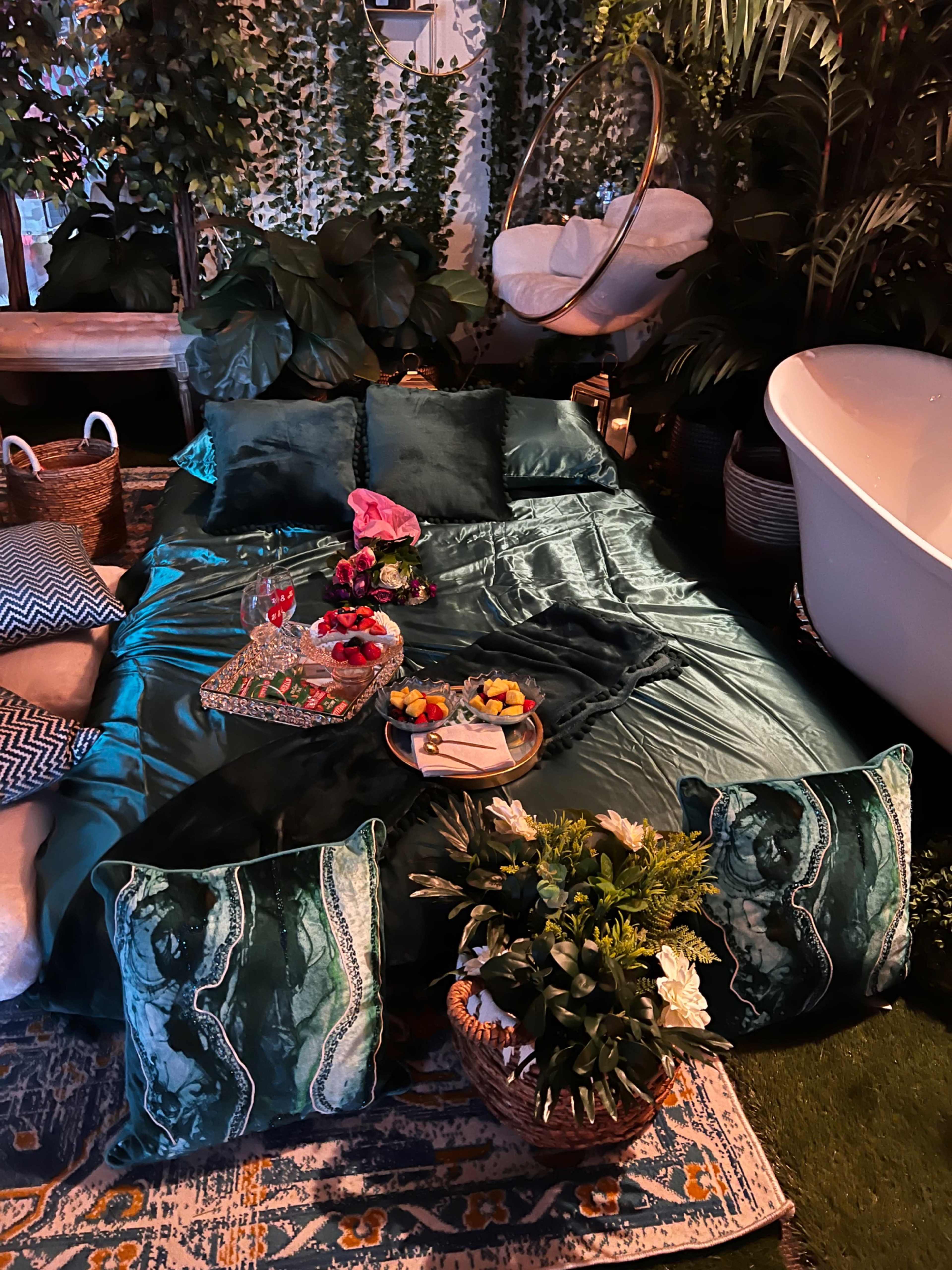 A cozy indoor setup with a green satin bedding arrangement, decorative pillows, a small table with snacks, and surrounded by various plants and a bathtub.