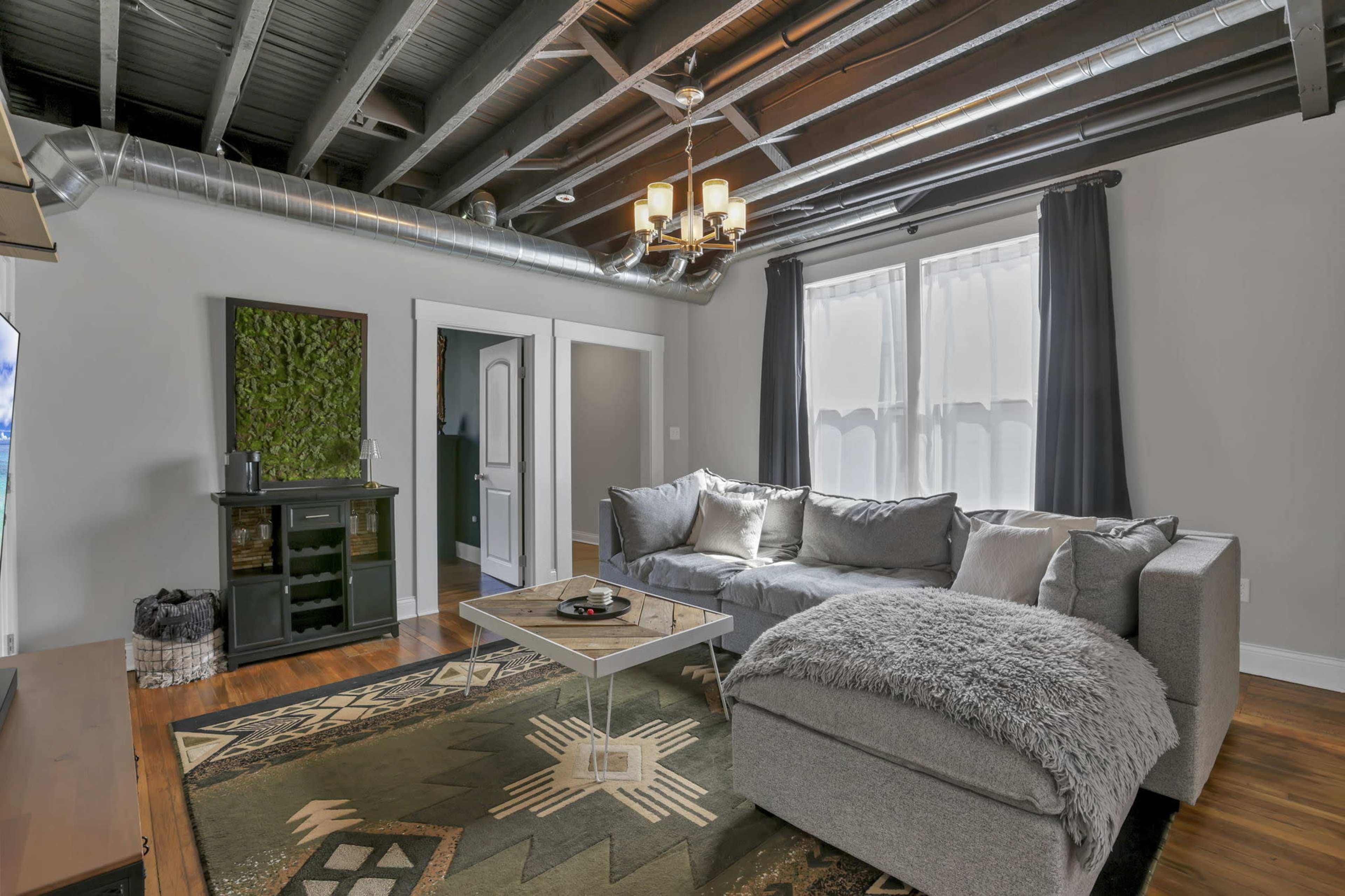The image shows a modern living room with a gray sectional sofa, a coffee table on an area rug, and exposed beams and ductwork on the ceiling.