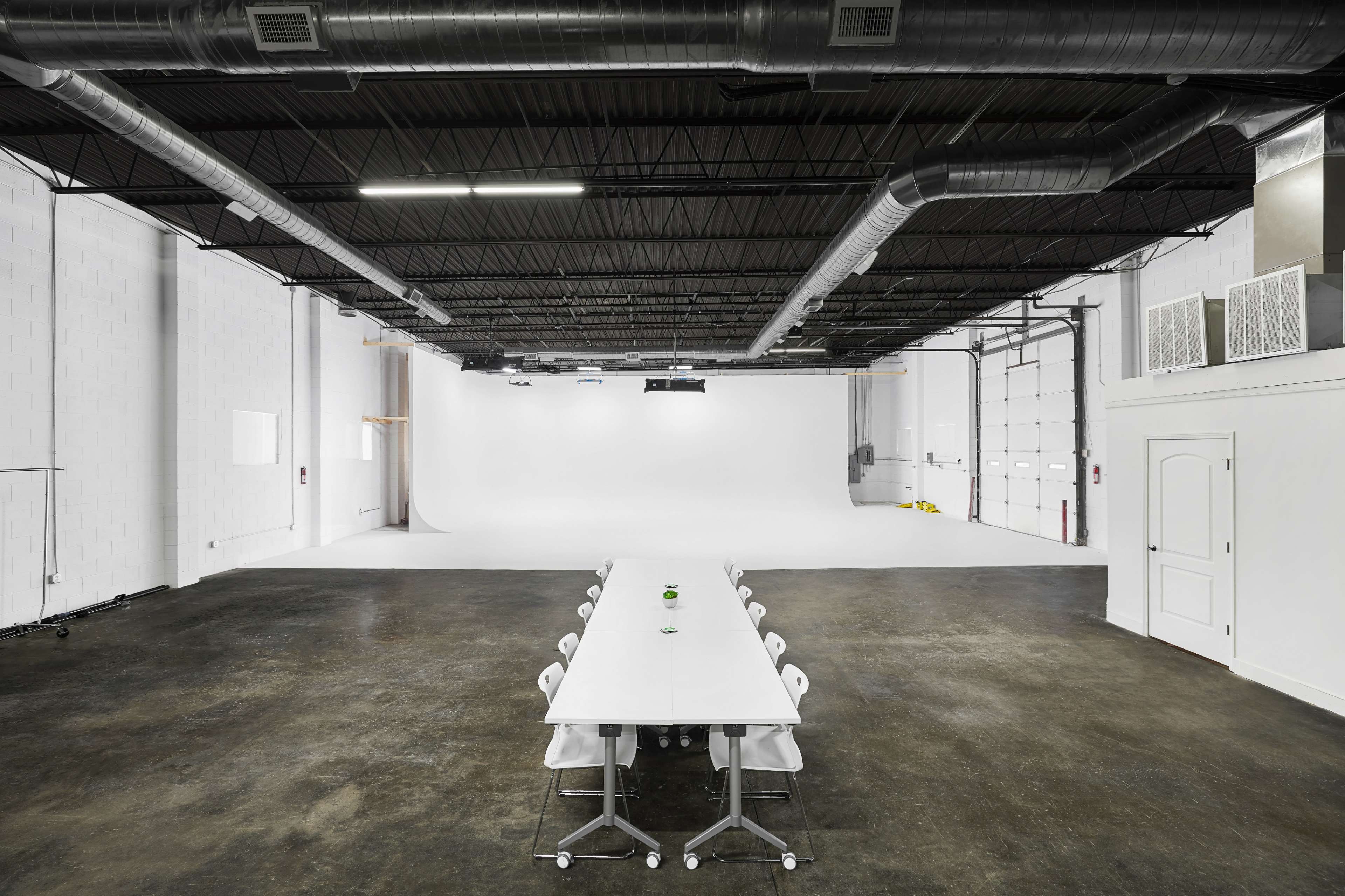 The image shows a large, empty studio space with a long white table surrounded by chairs, and a white backdrop at one end.