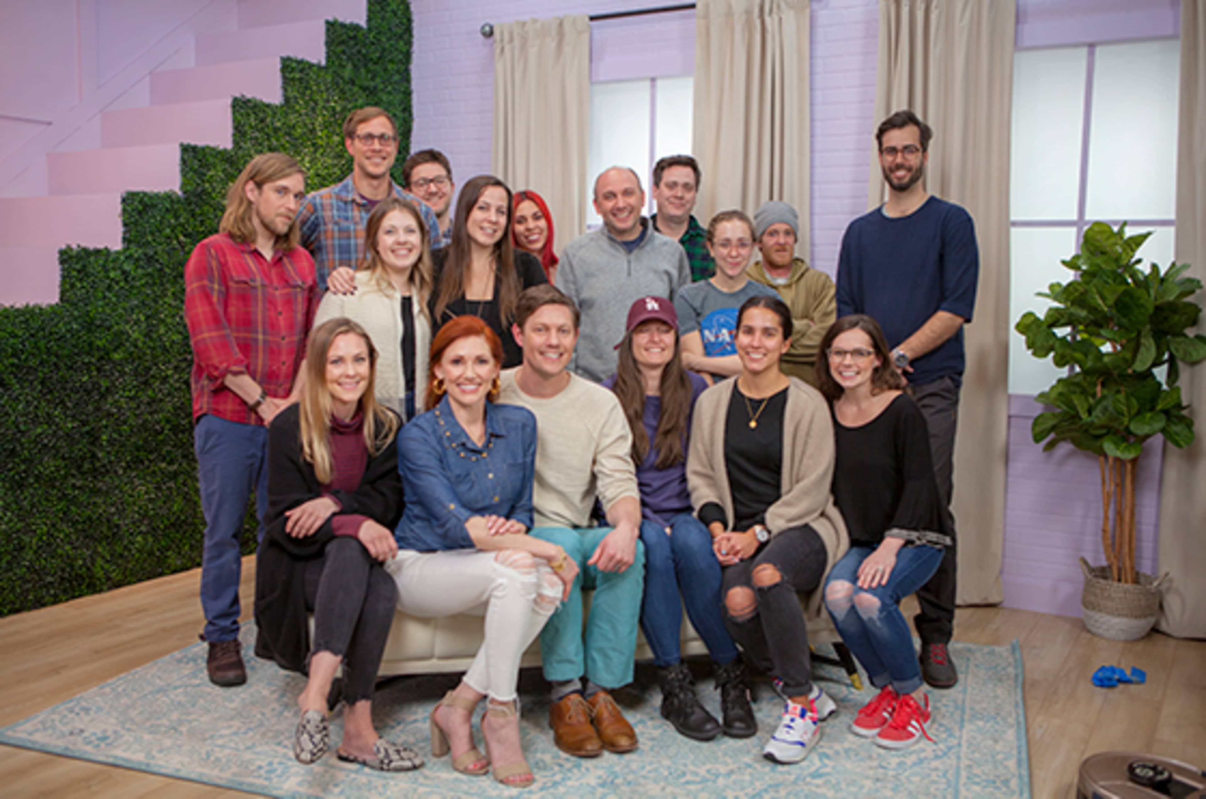A diverse group of people poses together in a bright room with a green plant and decorative backdrop.