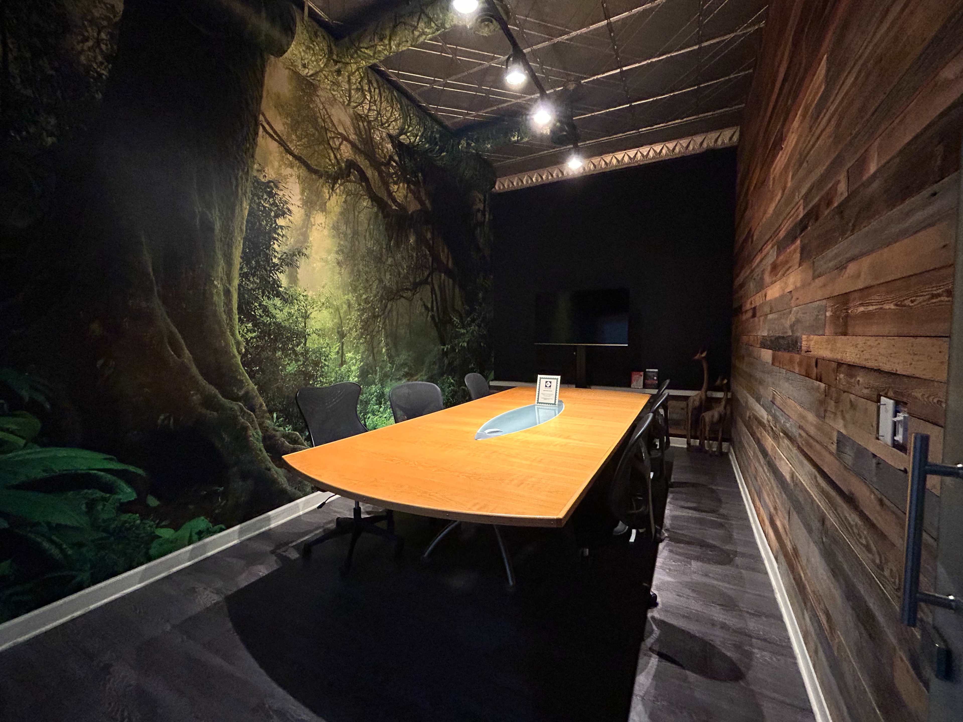 The image shows a modern conference room with a large wooden table, black chairs, and a mural of a forest on one wall.