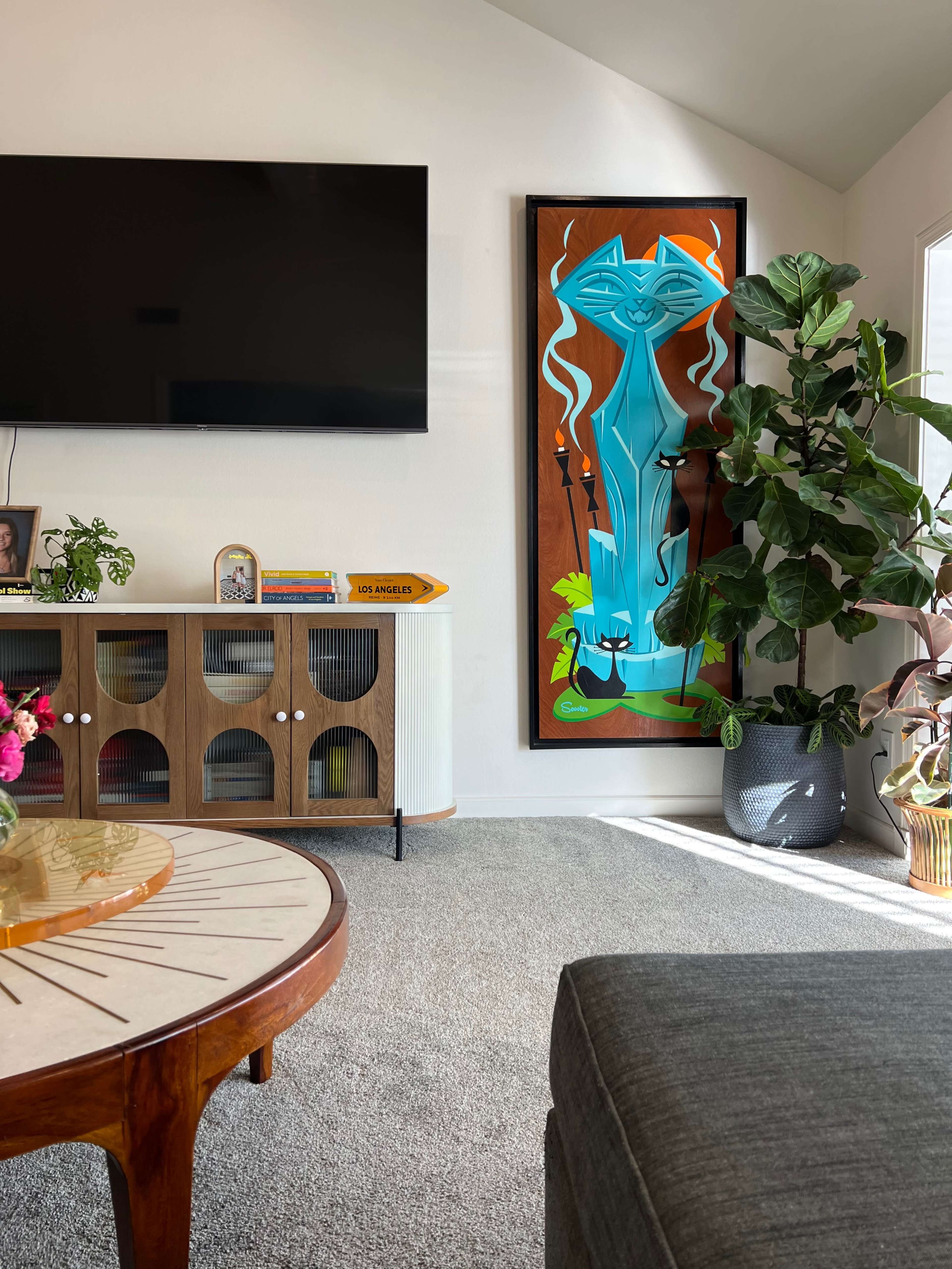 A living room with a large television, a decorative wall art piece of a blue and green cat-like figure, a potted plant, and a circular coffee table.