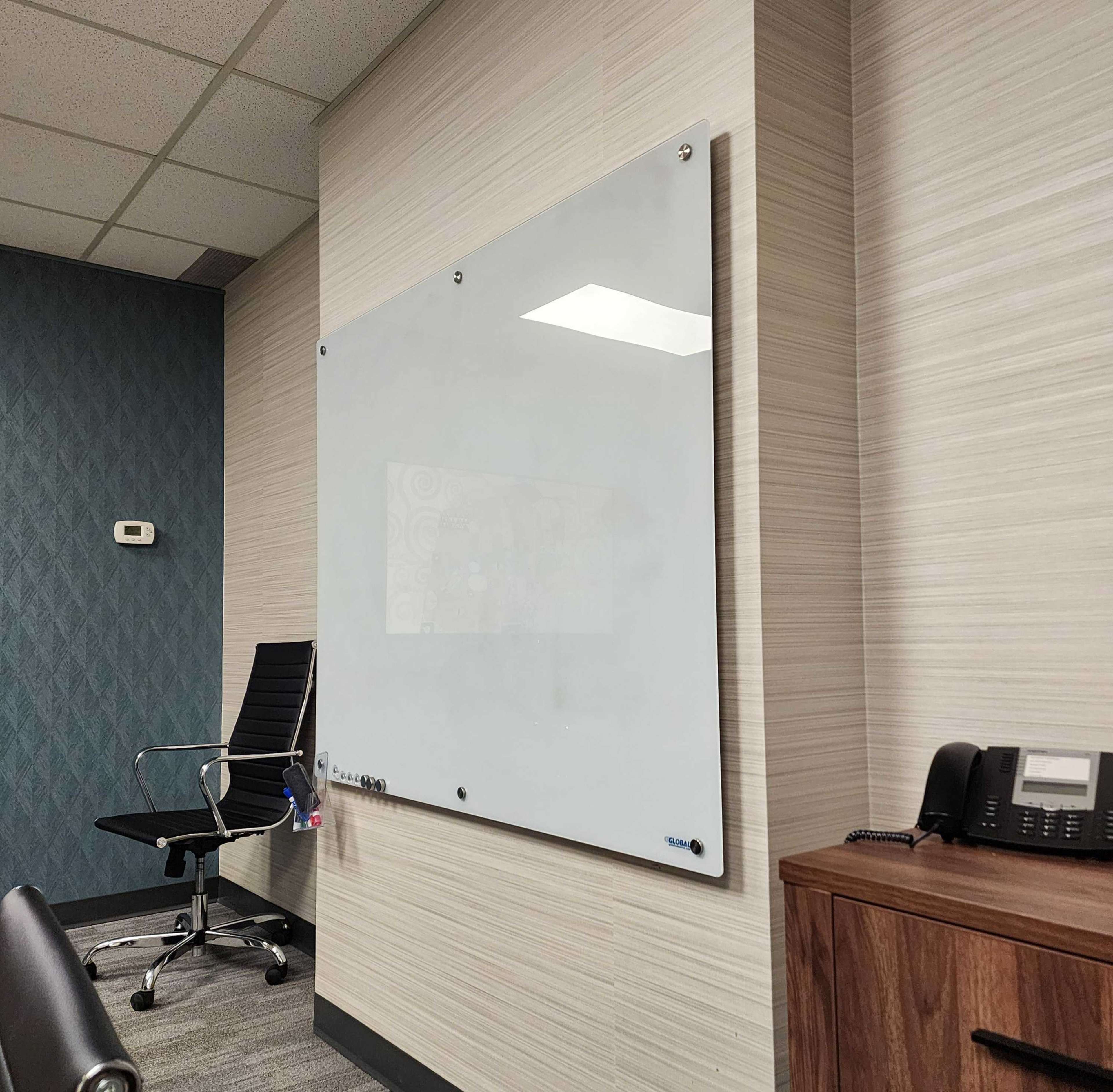 The image shows a modern office space featuring a large whiteboard mounted on a wall, a black office chair, and a telephone on a wooden desk.