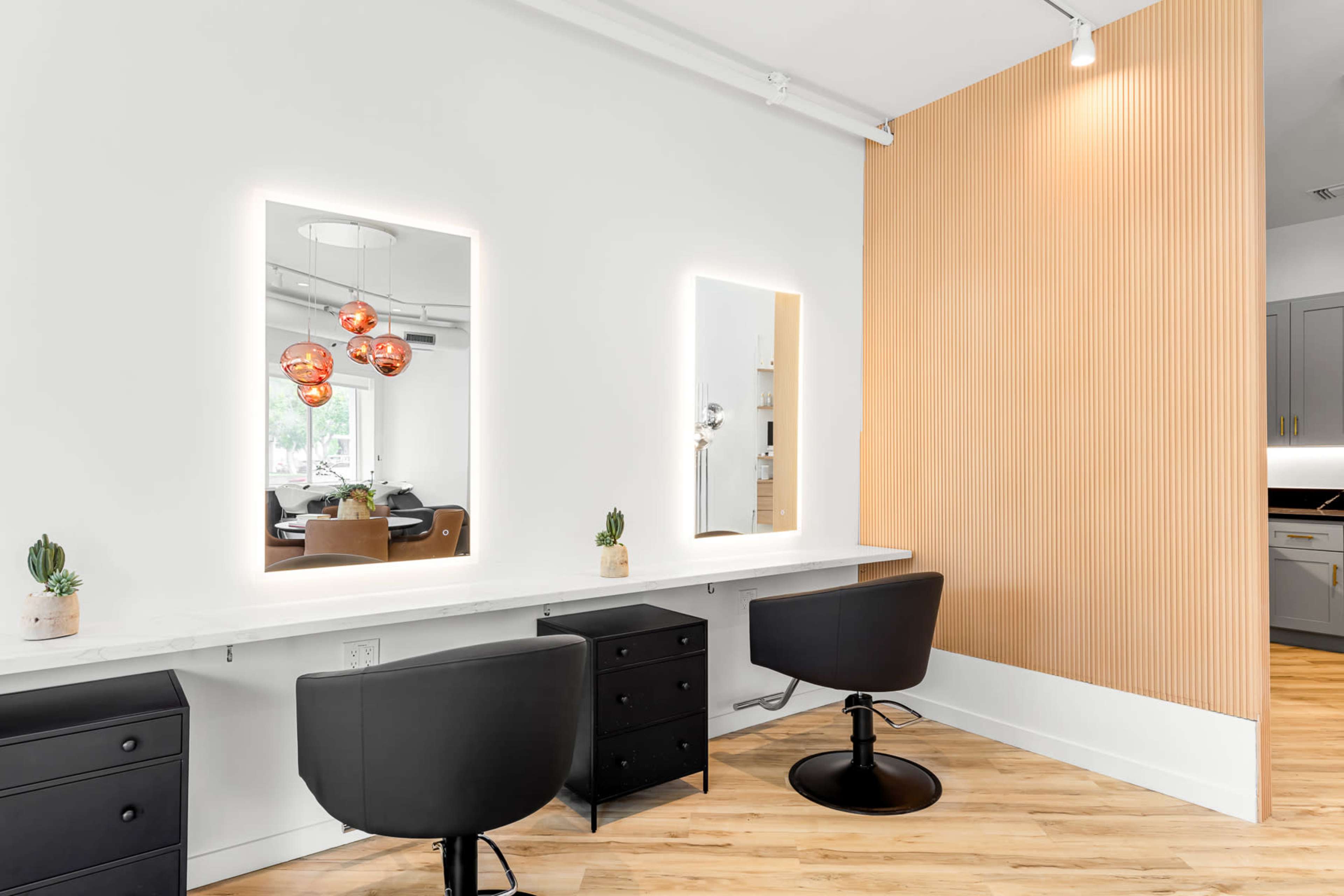 The image features a modern styling station with two black chairs, a marble countertop, and illuminated mirrors set against a wood-paneled wall.