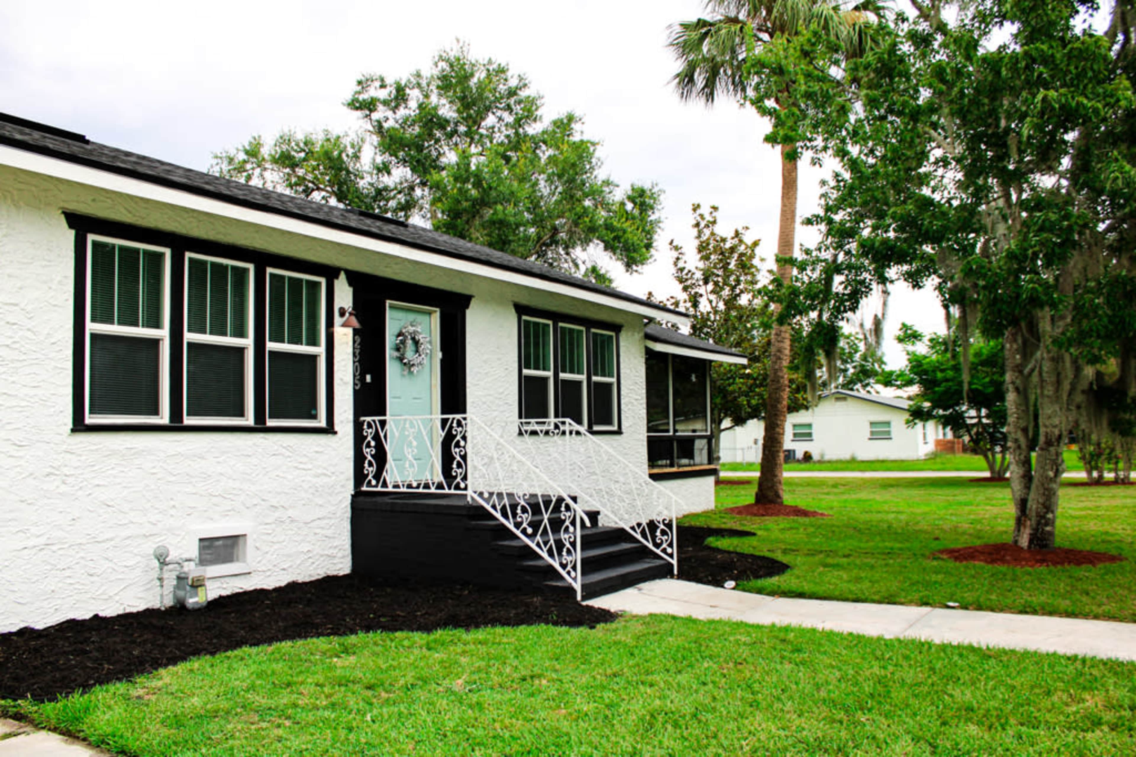 A single-story white house with a black front door and wrought iron railings is surrounded by green lawn and trees.