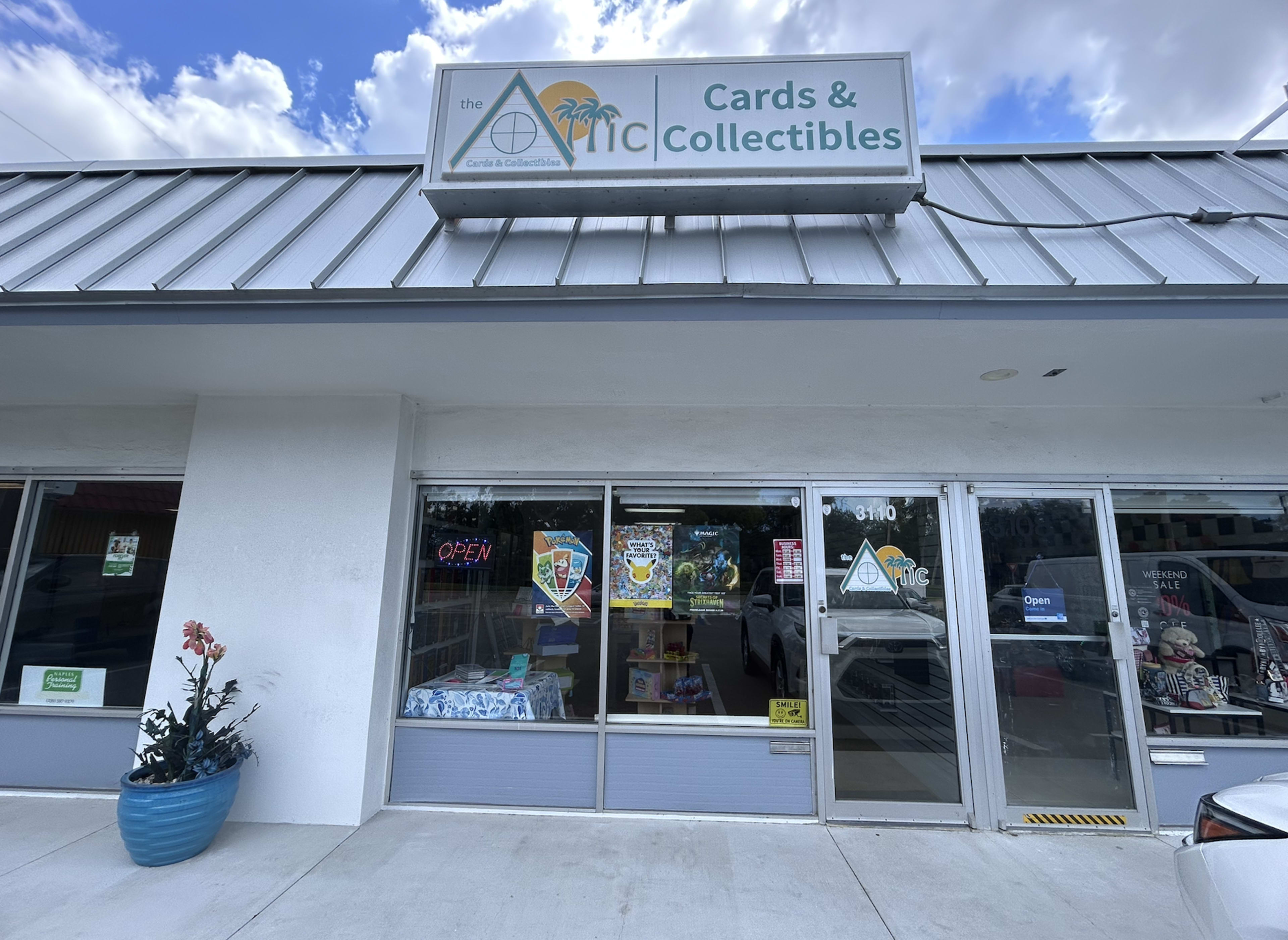 The image shows the exterior of a storefront named "The Attic Cards & Collectibles," featuring a blue plant pot and an "Open" sign in the window.