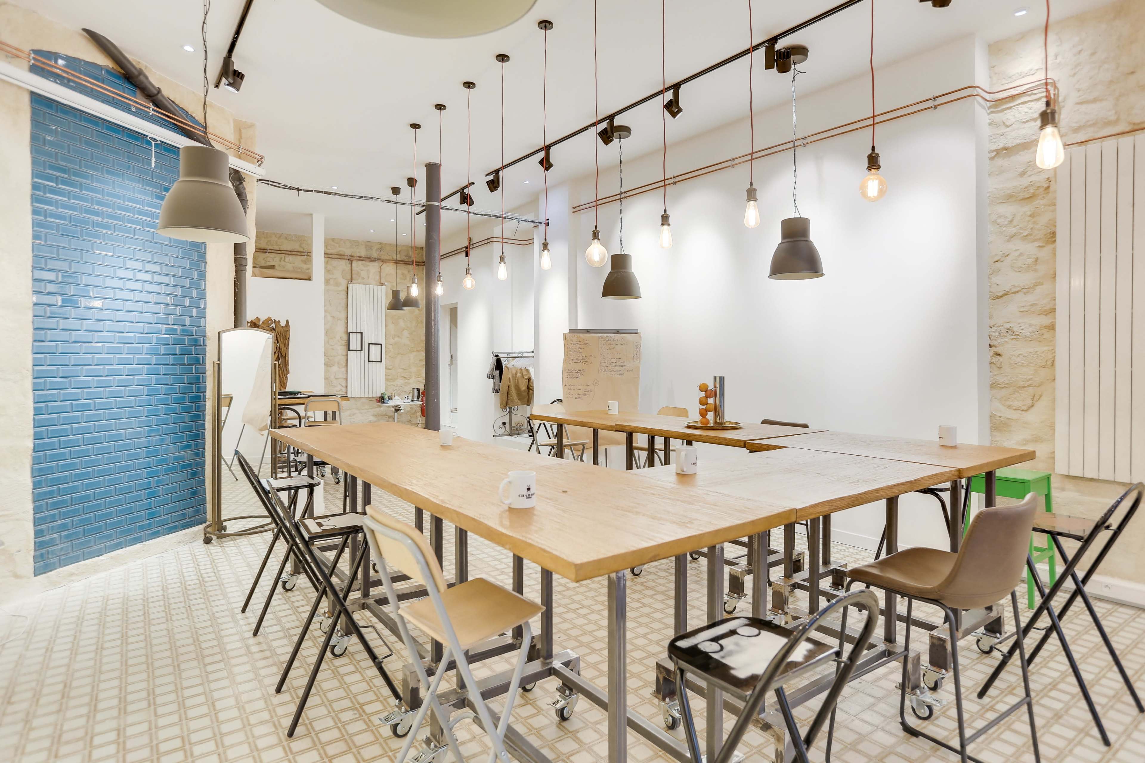 A spacious room features several long wooden tables surrounded by metal chairs, with pendant lights hanging from the ceiling and a blue-tiled wall on one side.