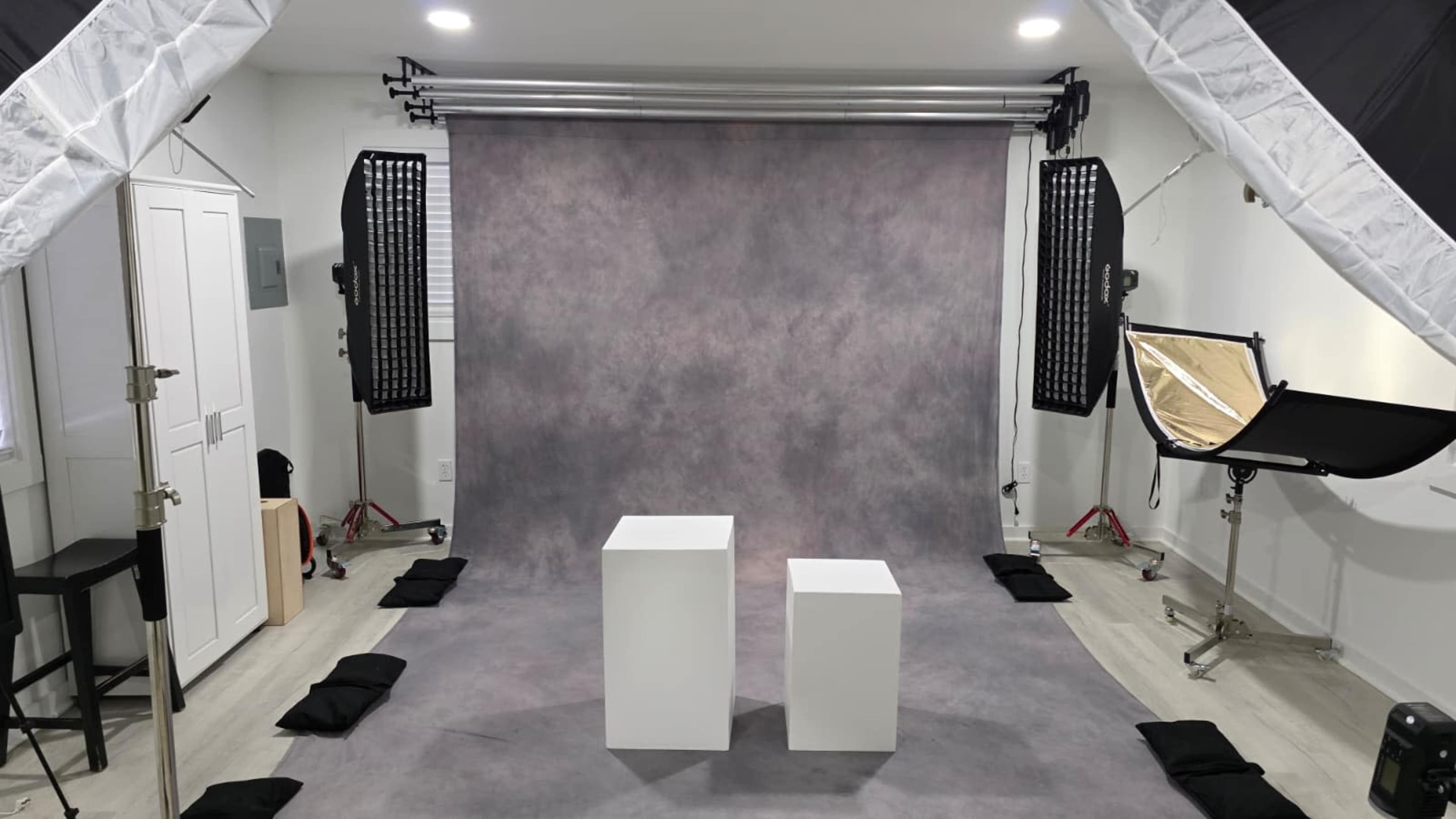 The image shows a photography studio featuring a gray backdrop, two white pedestals, and various lighting equipment arranged around the space.
