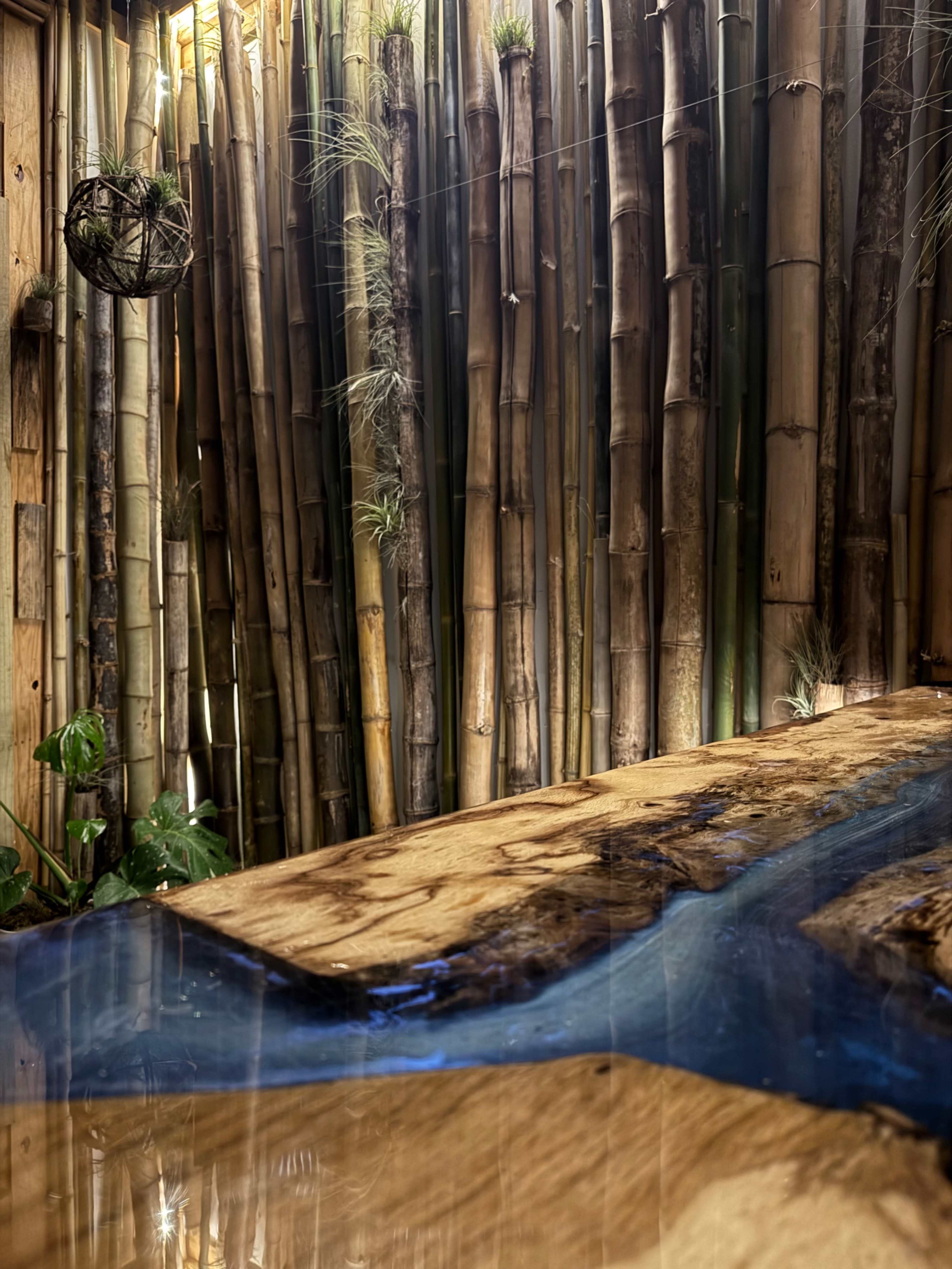 The image shows a natural interior space featuring tall bamboo stalks and a wooden table with a blue resin river design.