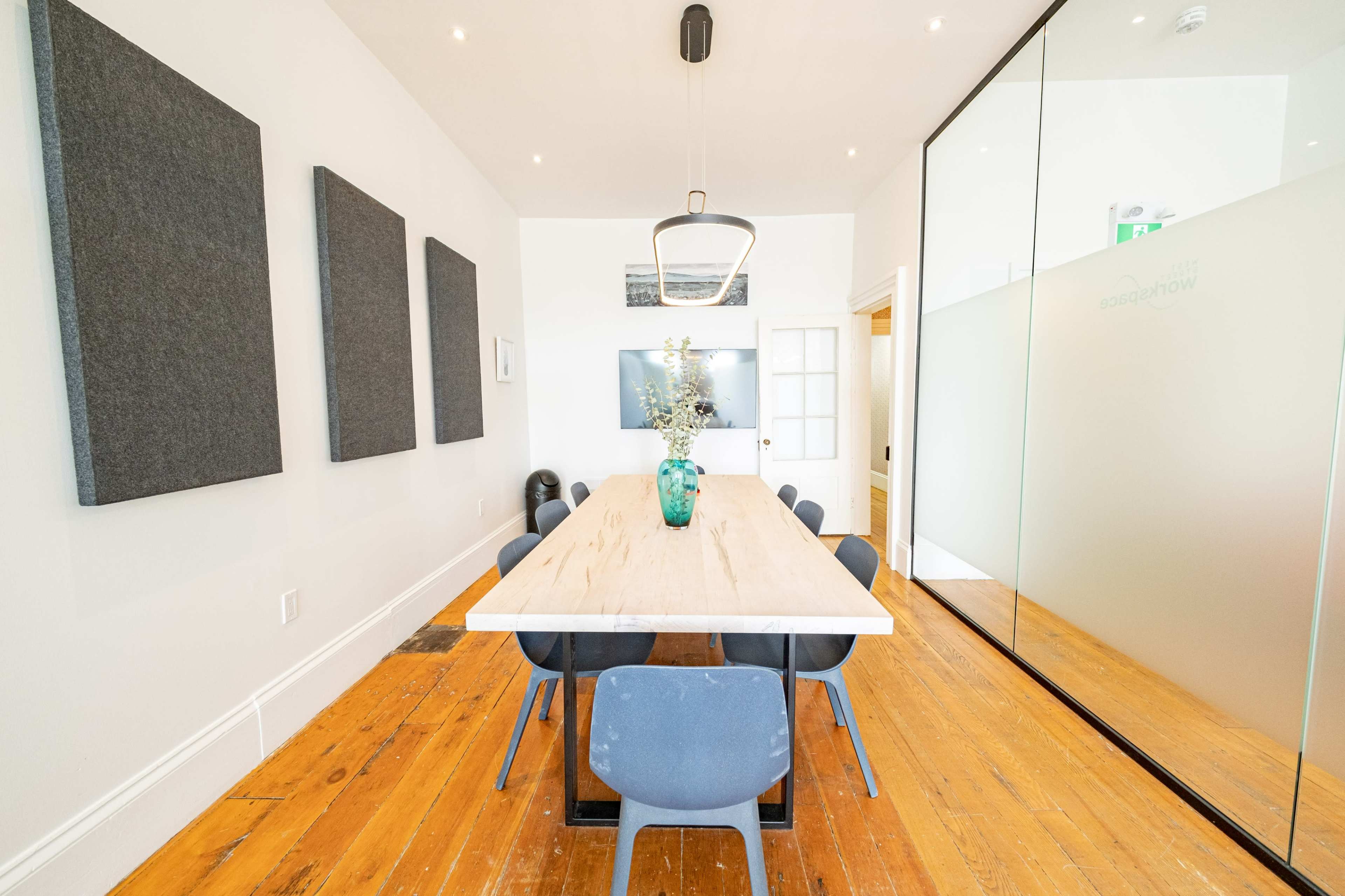 A long wooden table is surrounded by gray chairs in a well-lit meeting room with large wall panels and a glass partition.