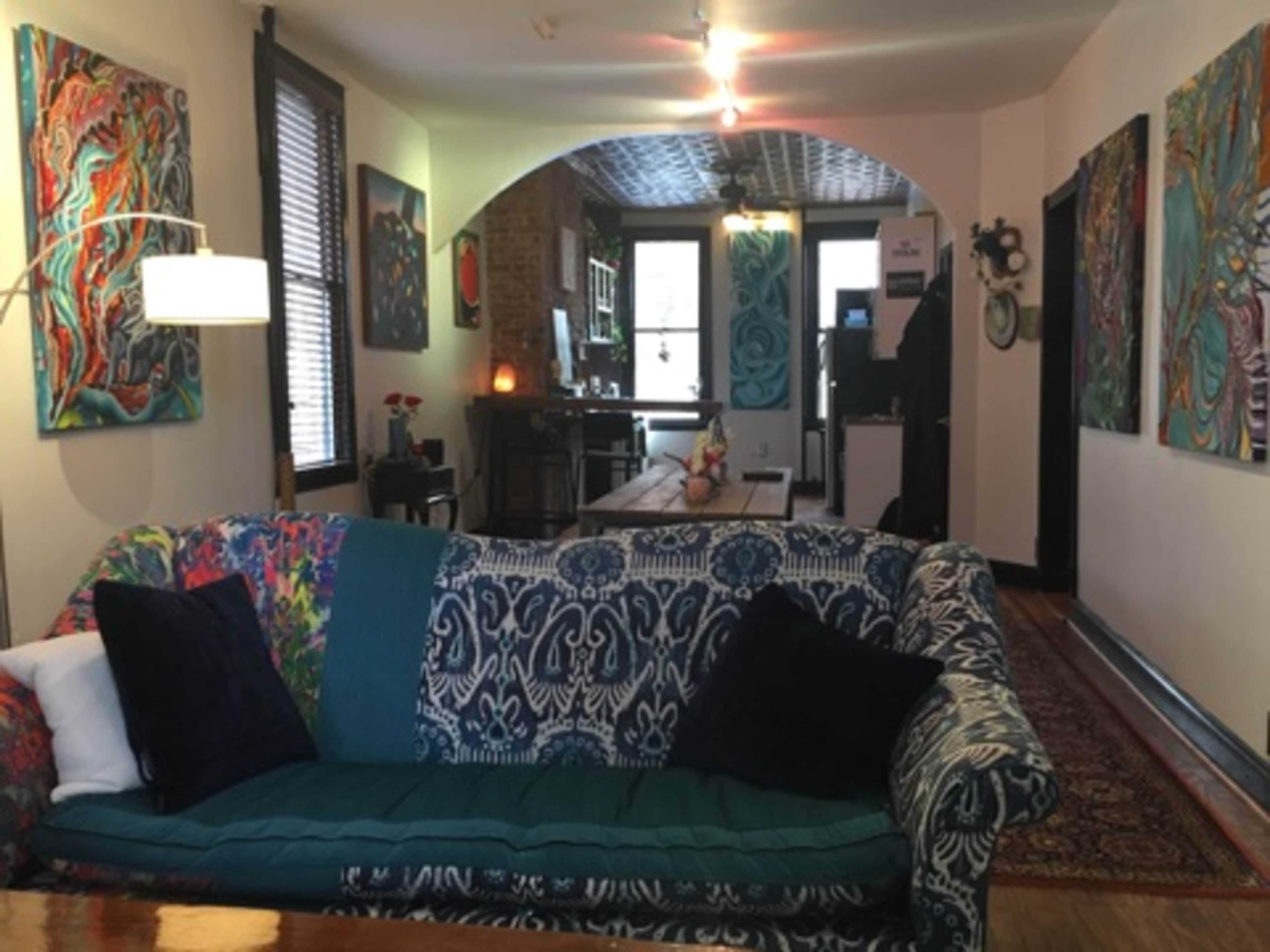 The image shows a living space featuring a patterned couch with blue and green tones, surrounded by colorful artwork on the walls and a wooden dining table in the background.
