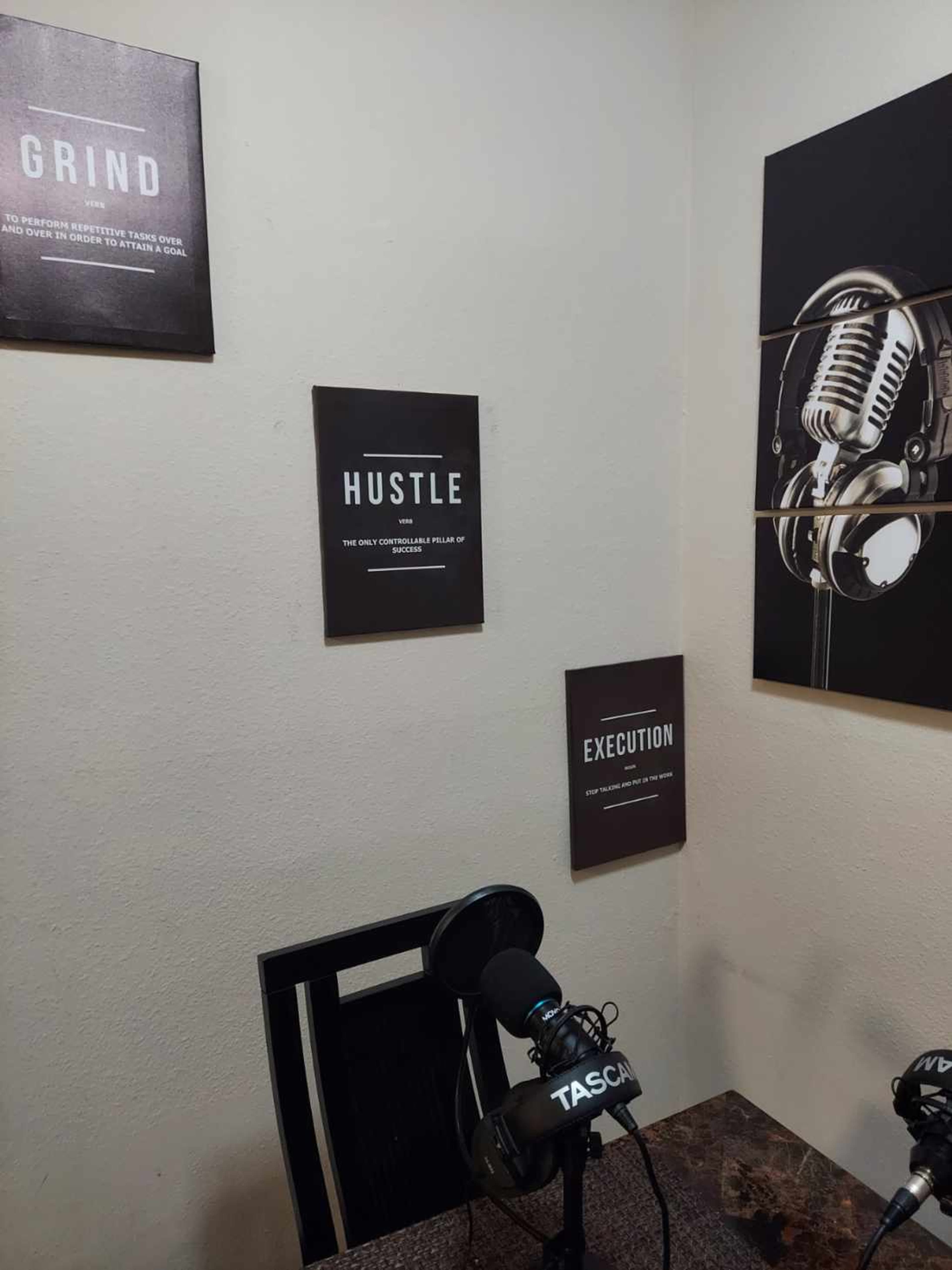 The image shows a small, motivational workspace with two posters displaying the words "GRIND," "HUSTLE," and "EXECUTION," alongside a microphone setup on a desk.