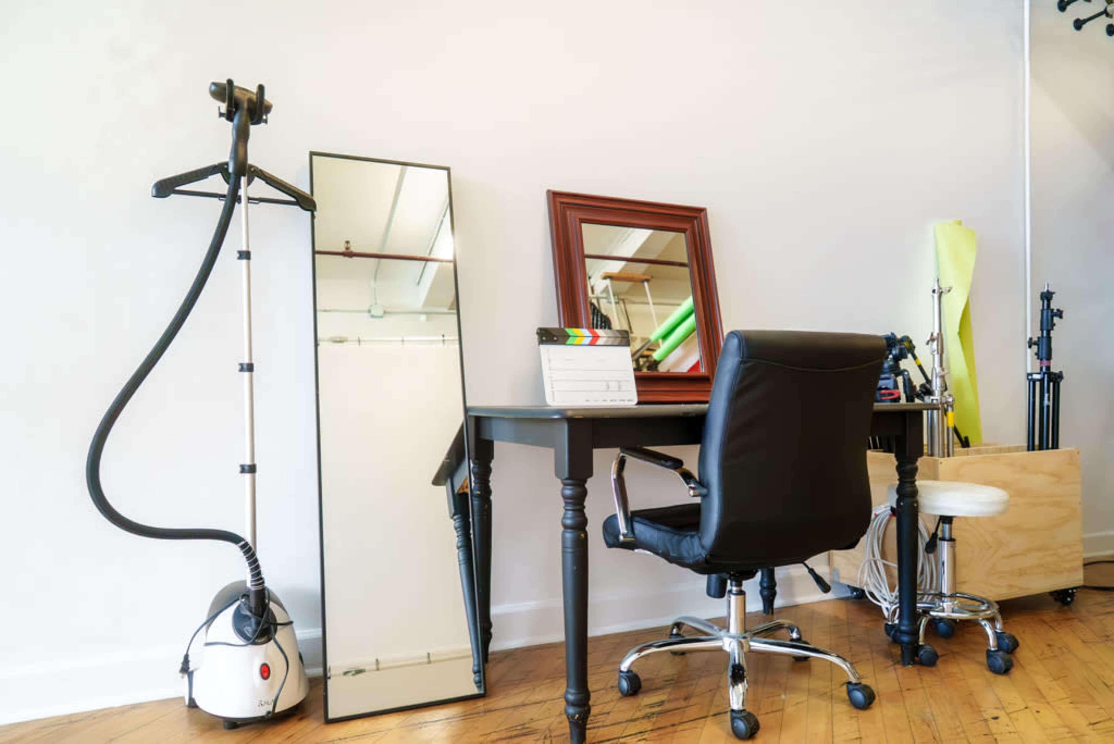 The image shows a minimalist workspace featuring a black desk, a chair, a full-length mirror, and a garment steamer beside a framed mirror.
