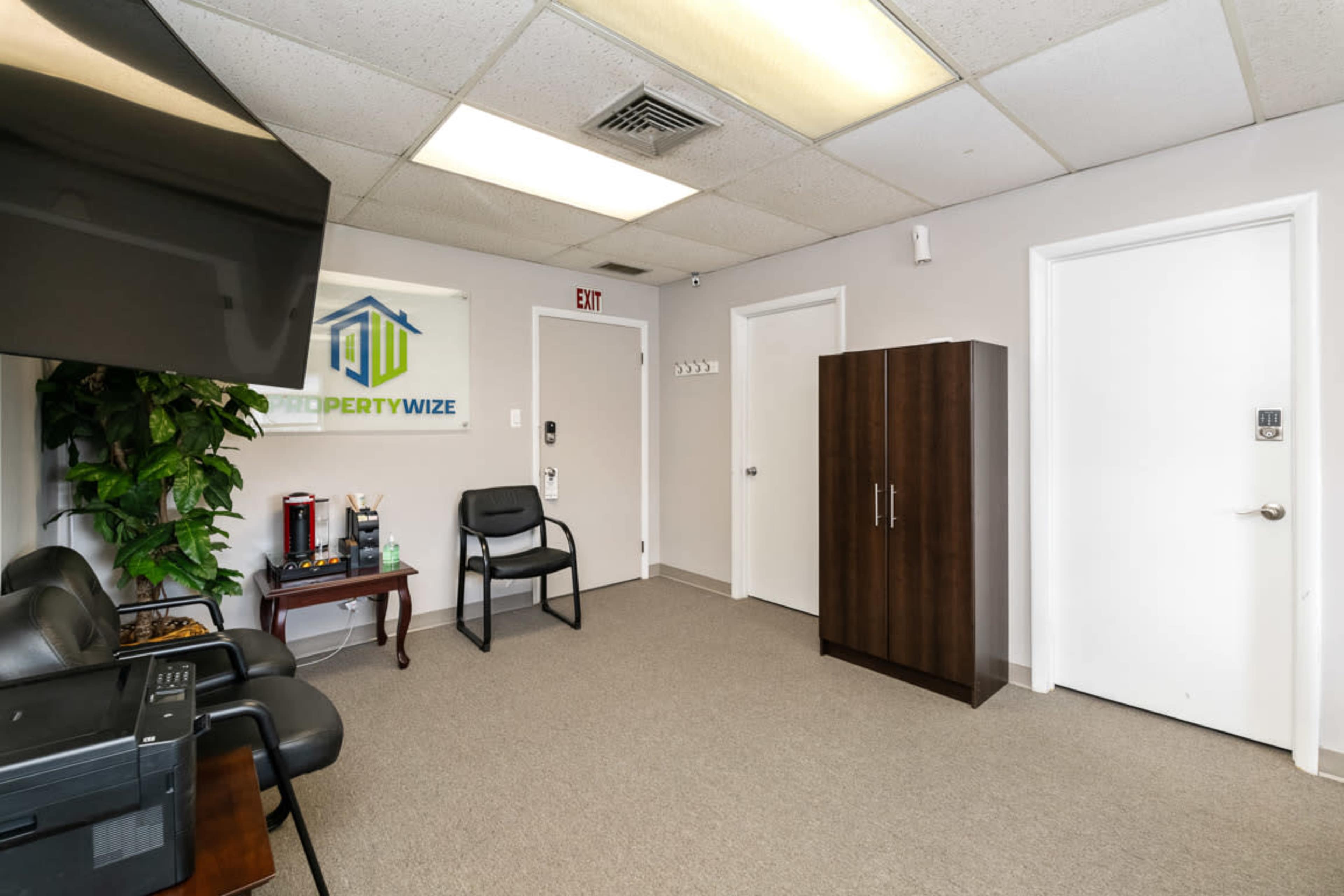 A small office space with a television on a stand, a chair, a cabinet, and a plant, featuring a door and a wall with a logo.