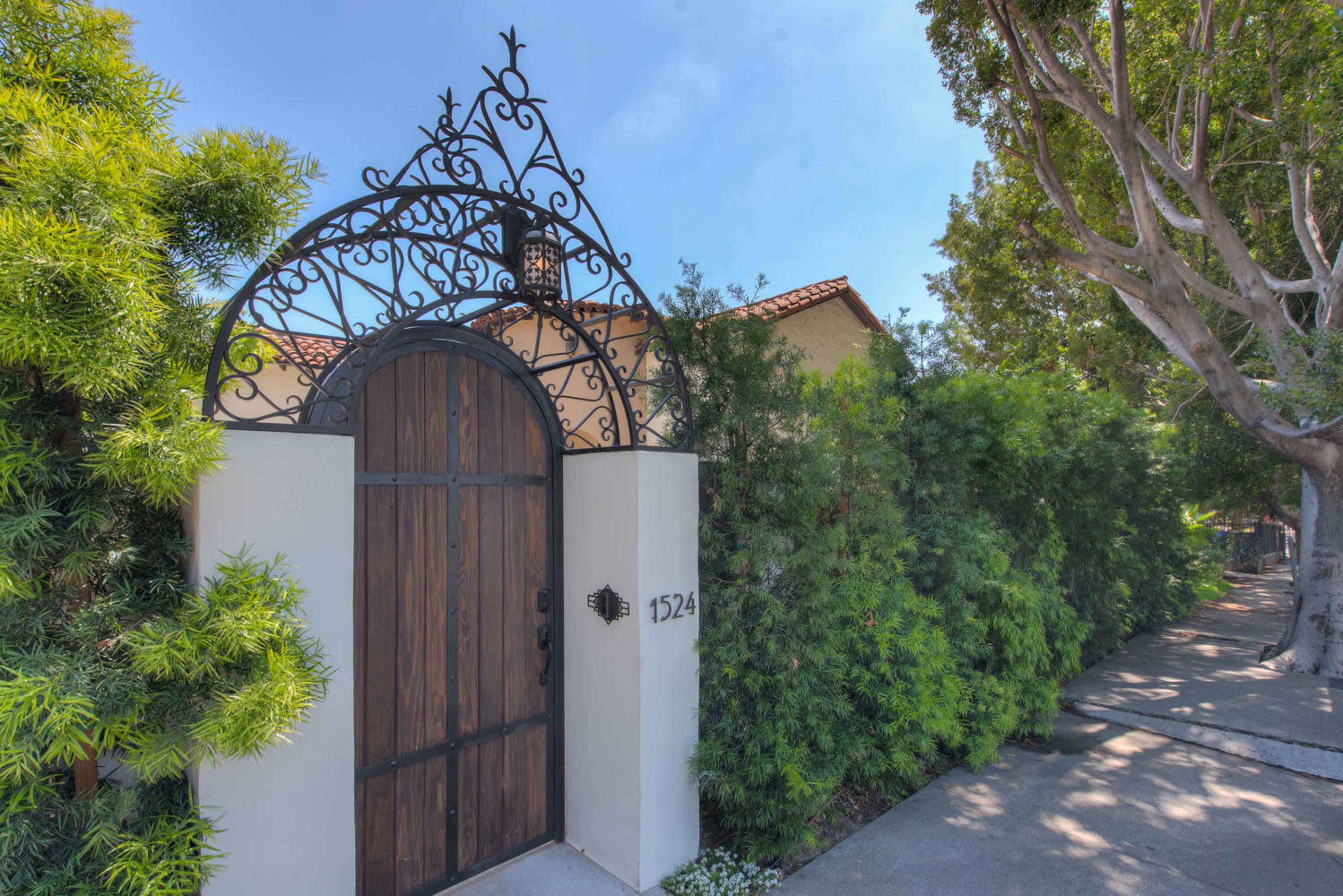 A decorative wrought iron gate with a wooden door, framed by lush greenery and a concrete pathway.