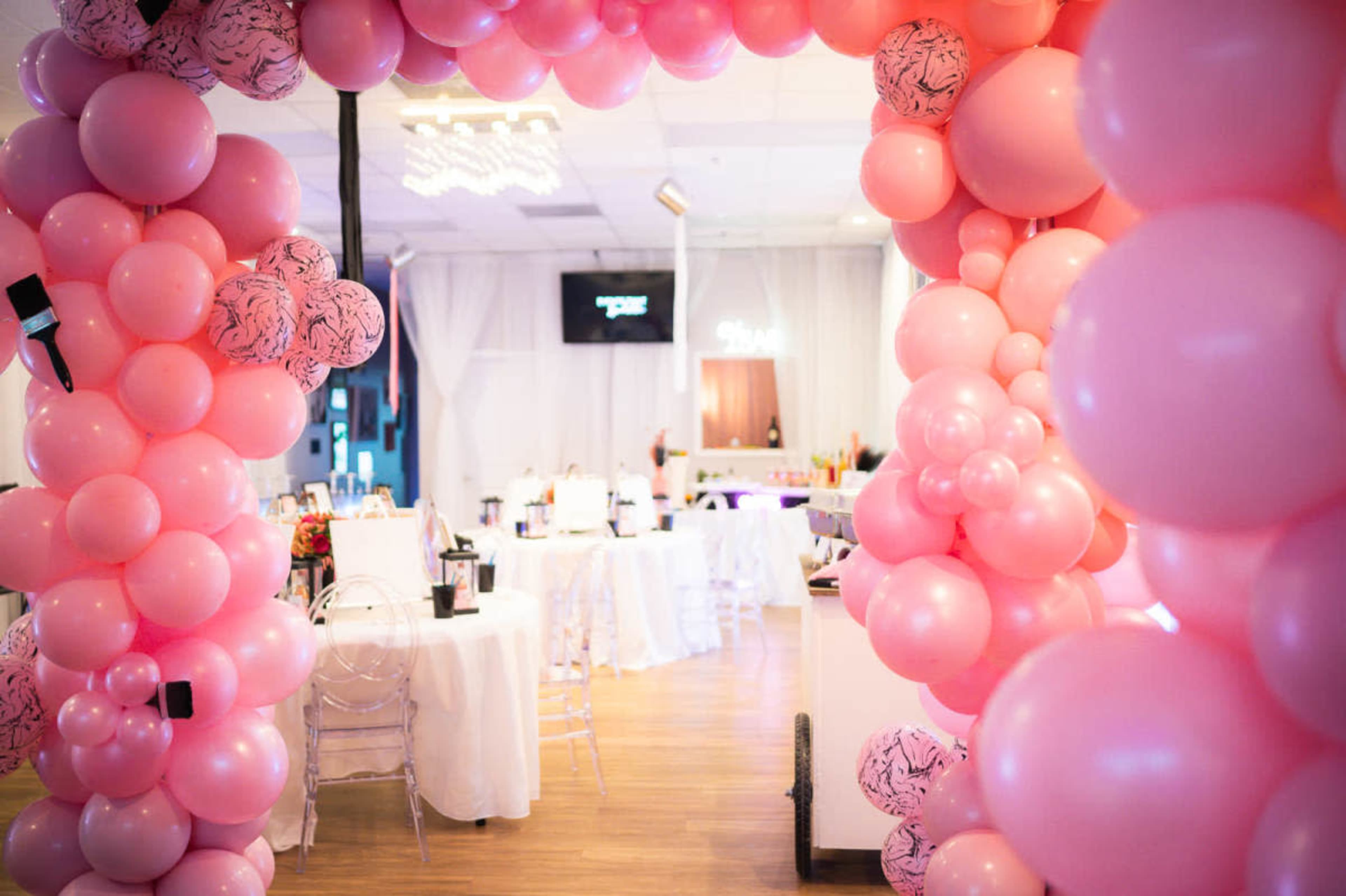A large arch made of pink balloons frames a decorated event space featuring tables and a bar.