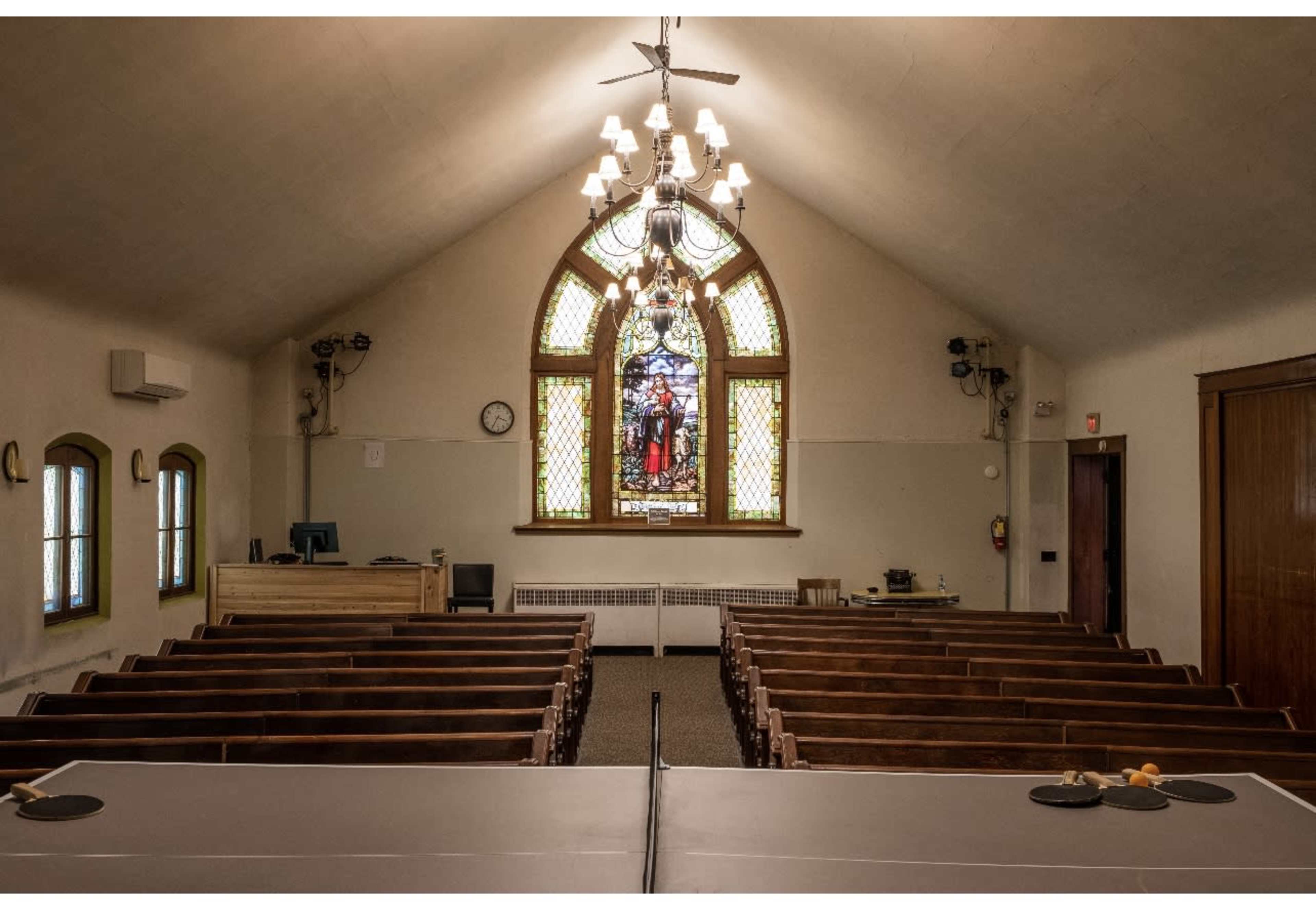 An interior view of a church, featuring wooden pews, a stained glass window, and a chandelier.