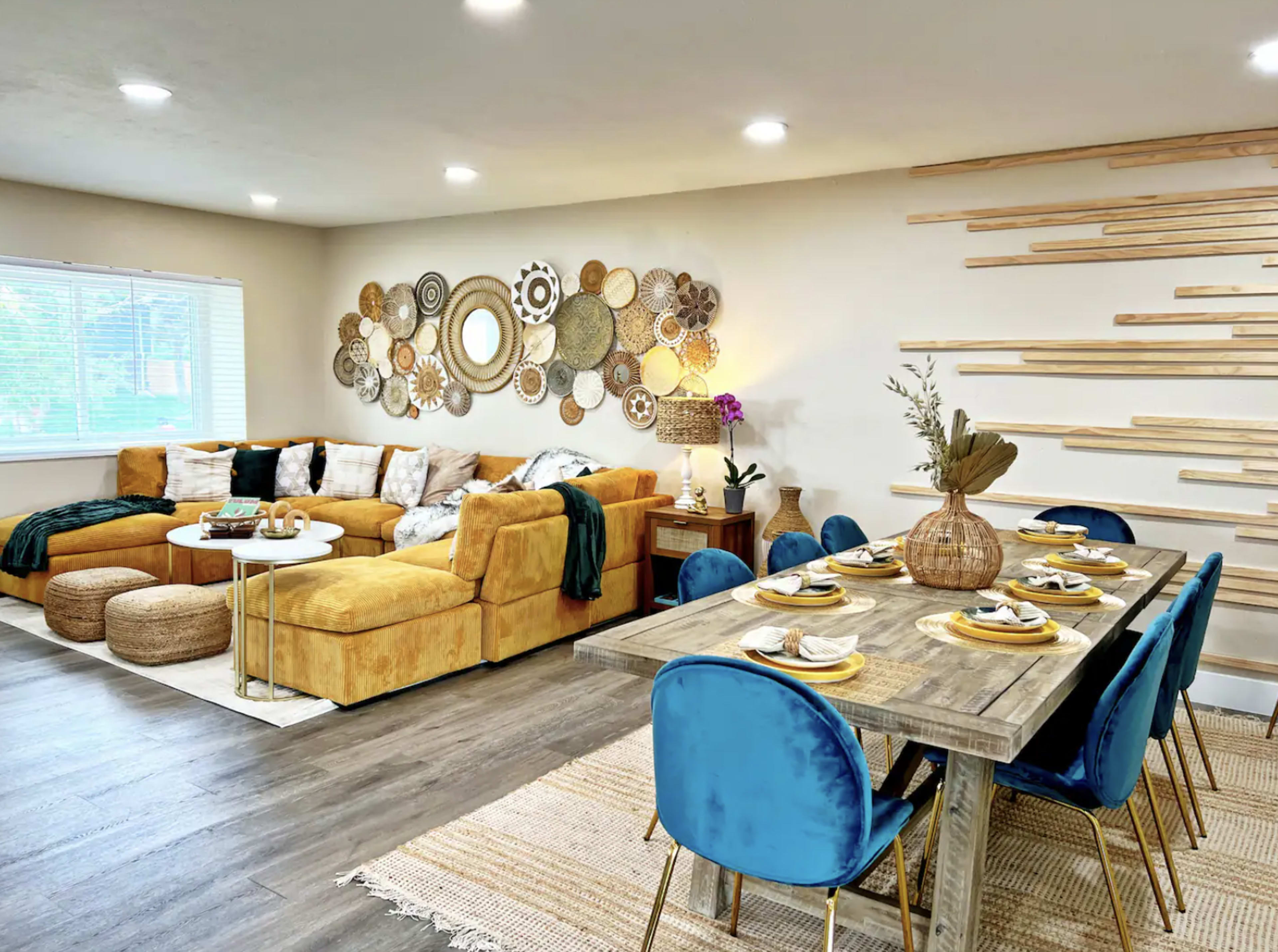 A spacious living and dining area features a yellow sectional sofa, a wooden dining table with blue chairs, and decorative wall art made of natural materials.