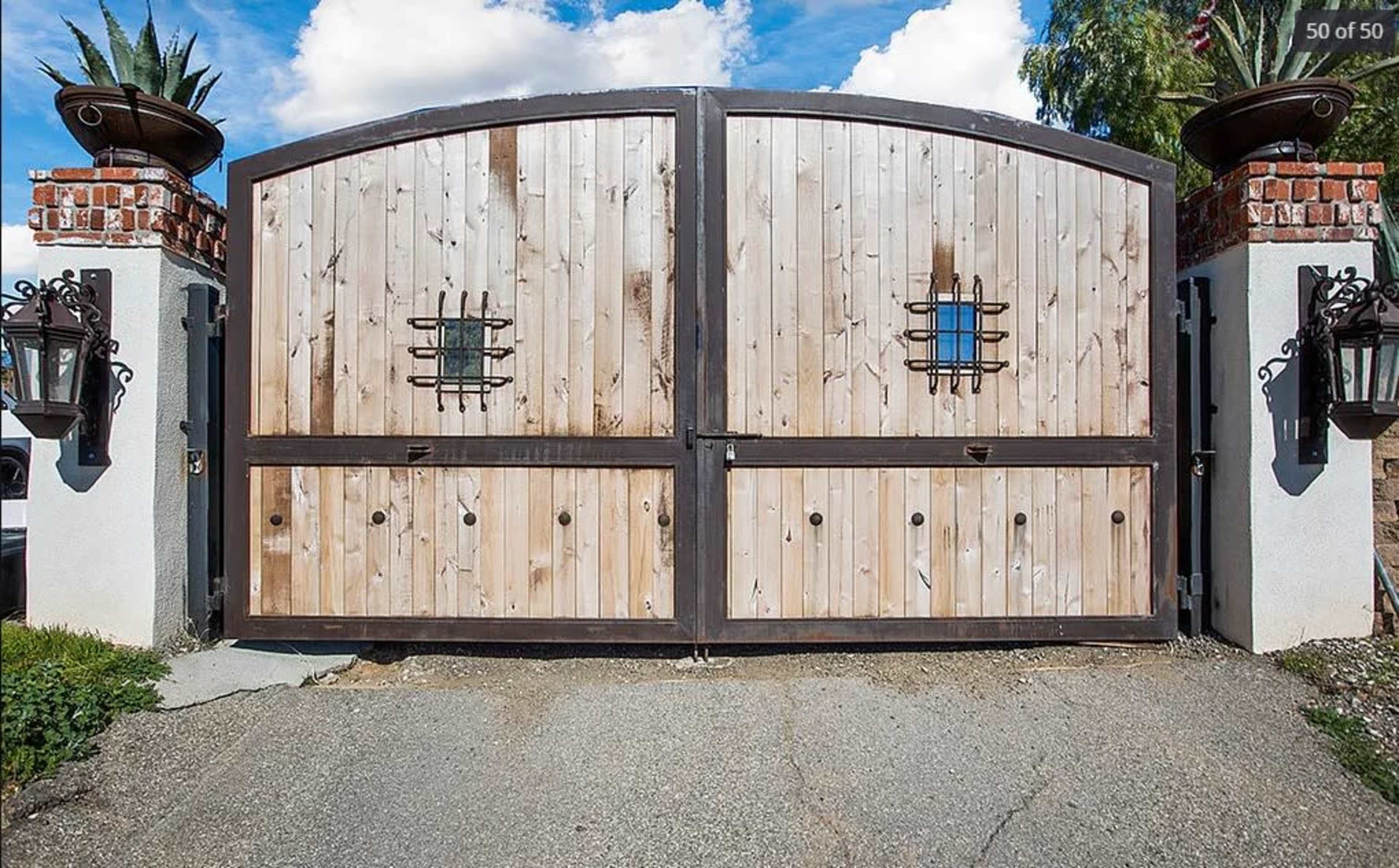 A large wooden double gate with metal accents stands closed at the entrance, flanked by brick pillars and decorative lanterns.