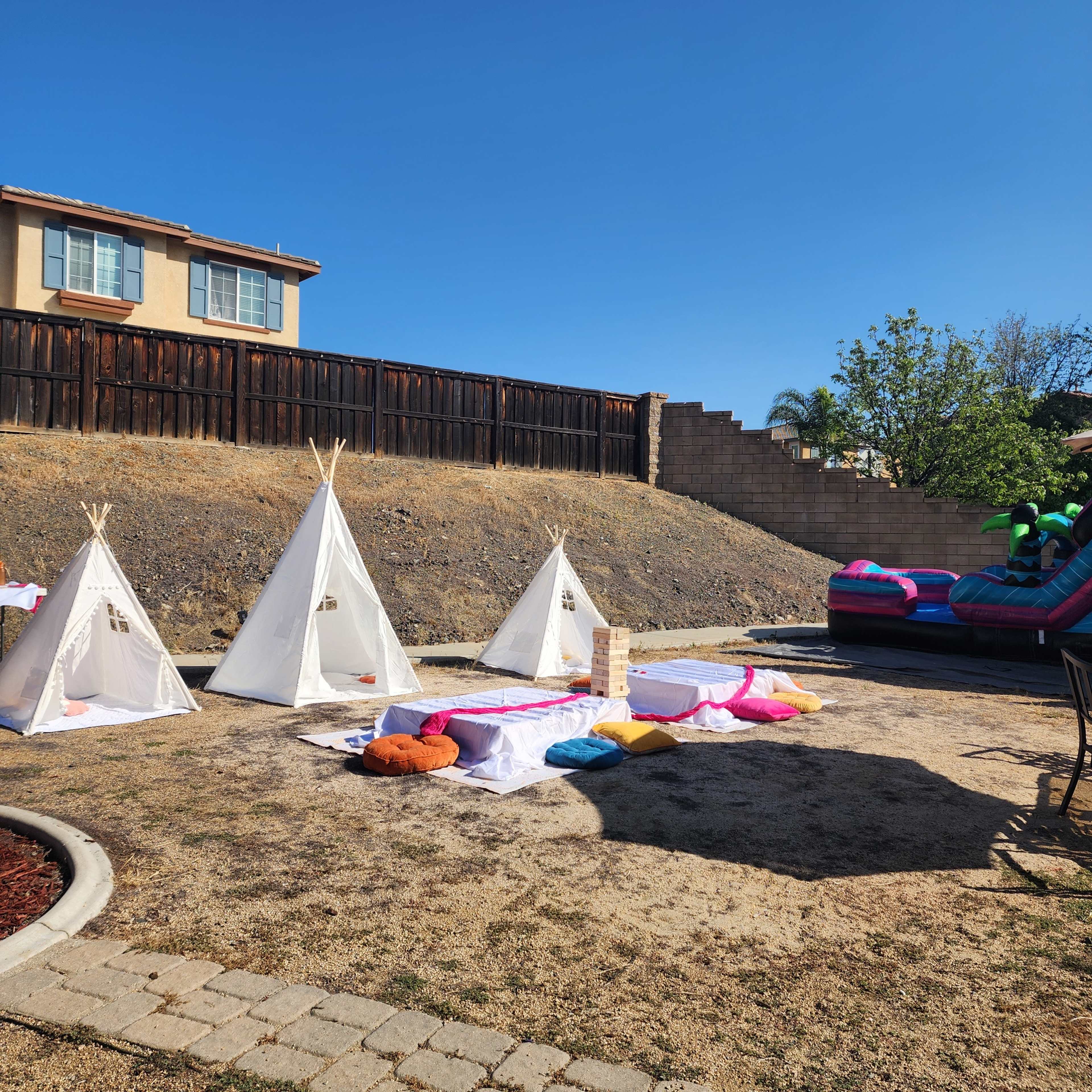 The image shows a backyard with three white teepees, a colorful blanket area, a large inflatable slide, and a wooden fence in the background.