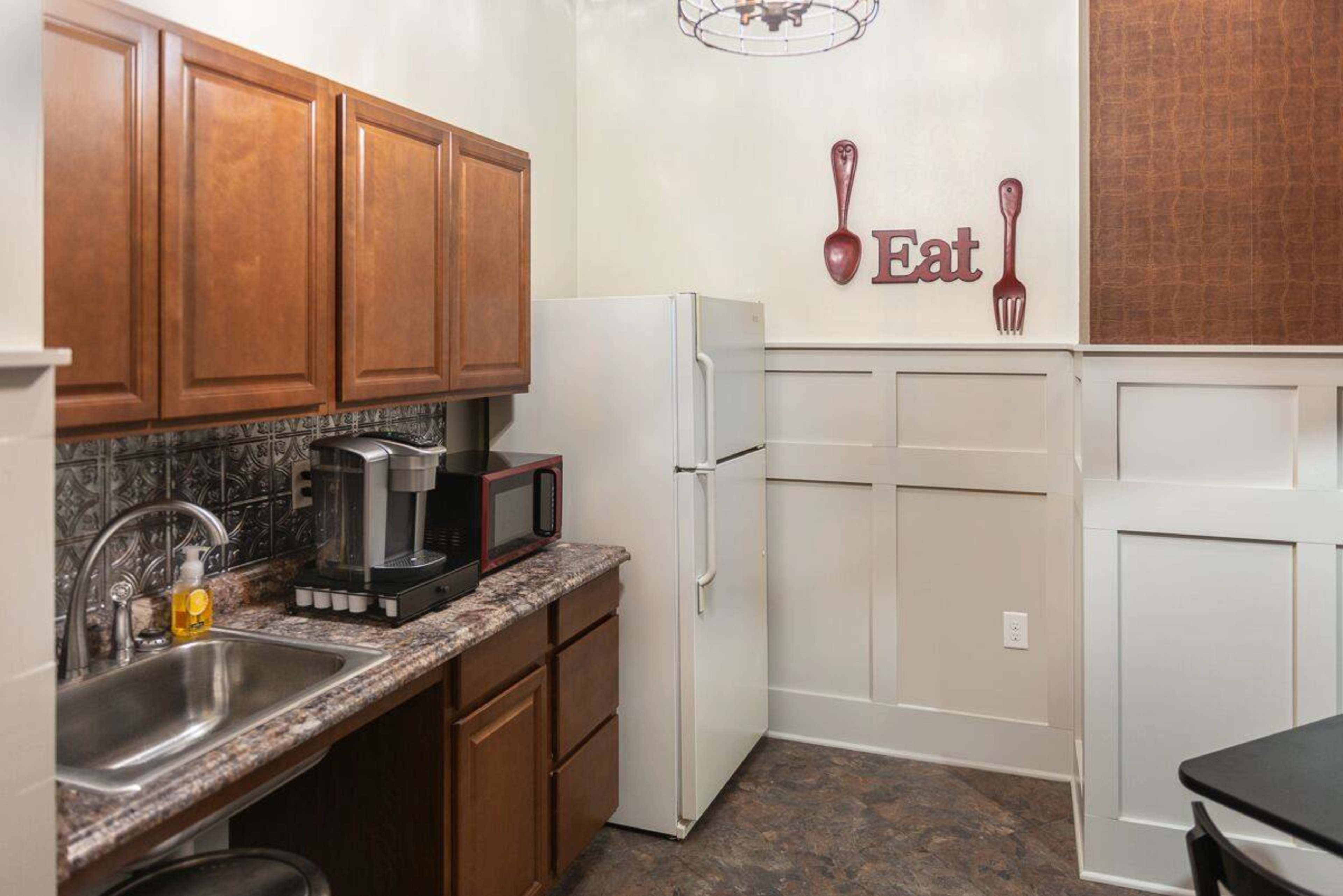 A small kitchen area with wooden cabinets, a white refrigerator, a sink, and a coffee maker, along with a wall decor that reads "Eat."