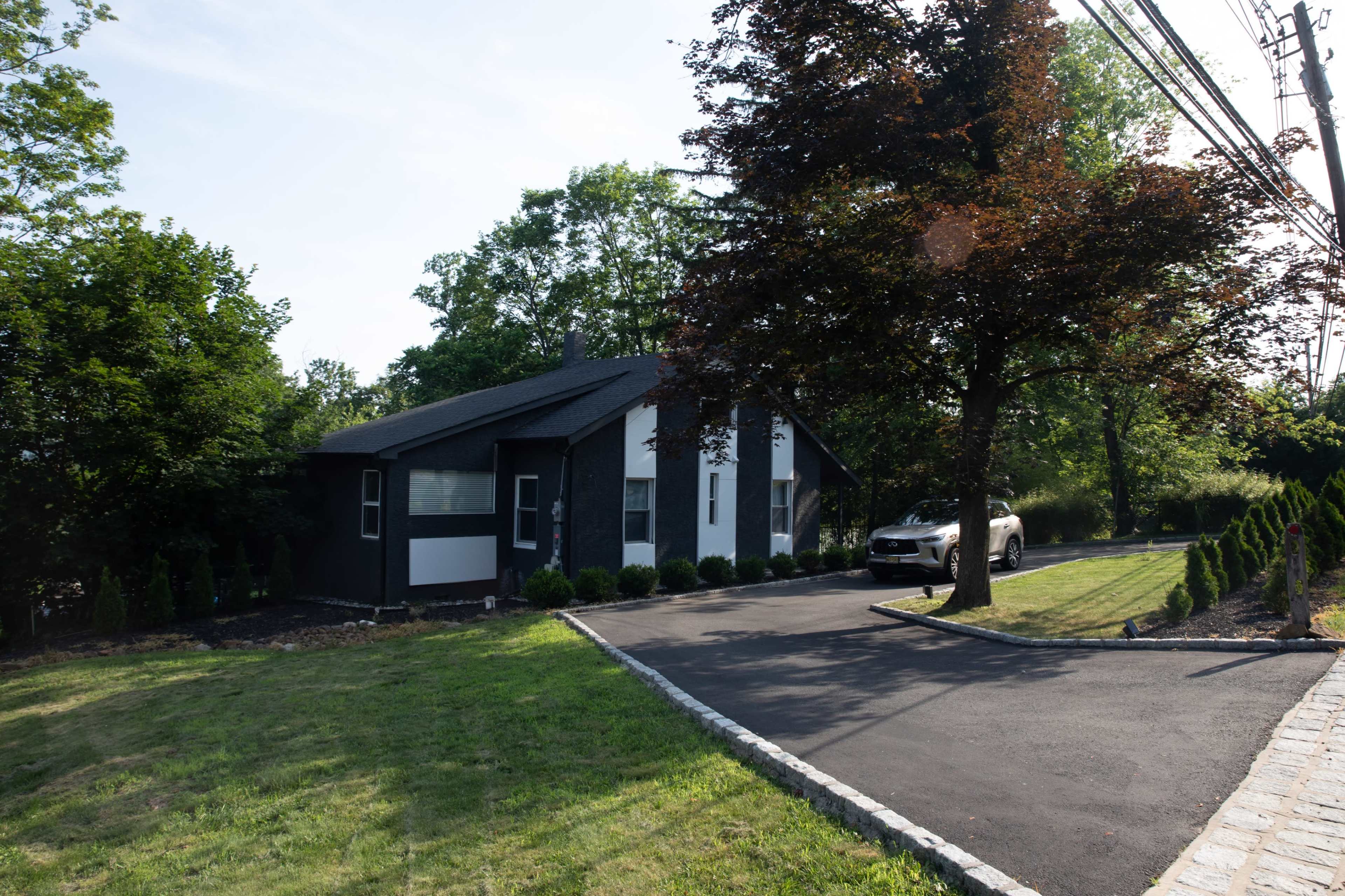 A modern, black-and-white house is positioned at the end of a paved driveway lined with small bushes, shaded by a tree, with a car parked nearby.