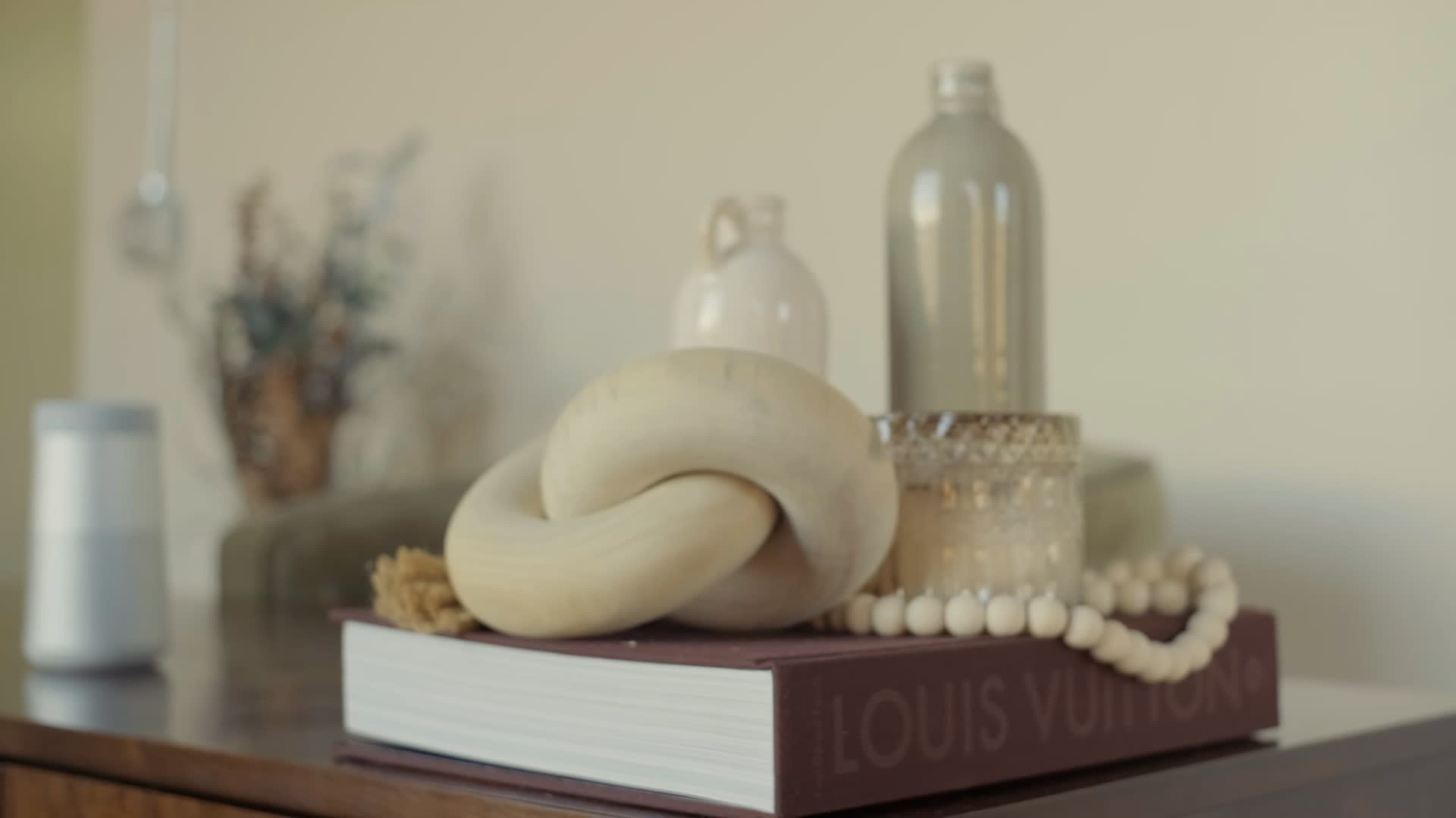 A wooden knot sculpture rests on a Louis Vuitton book beside a glass bottle and a decorative candle on a wooden table.