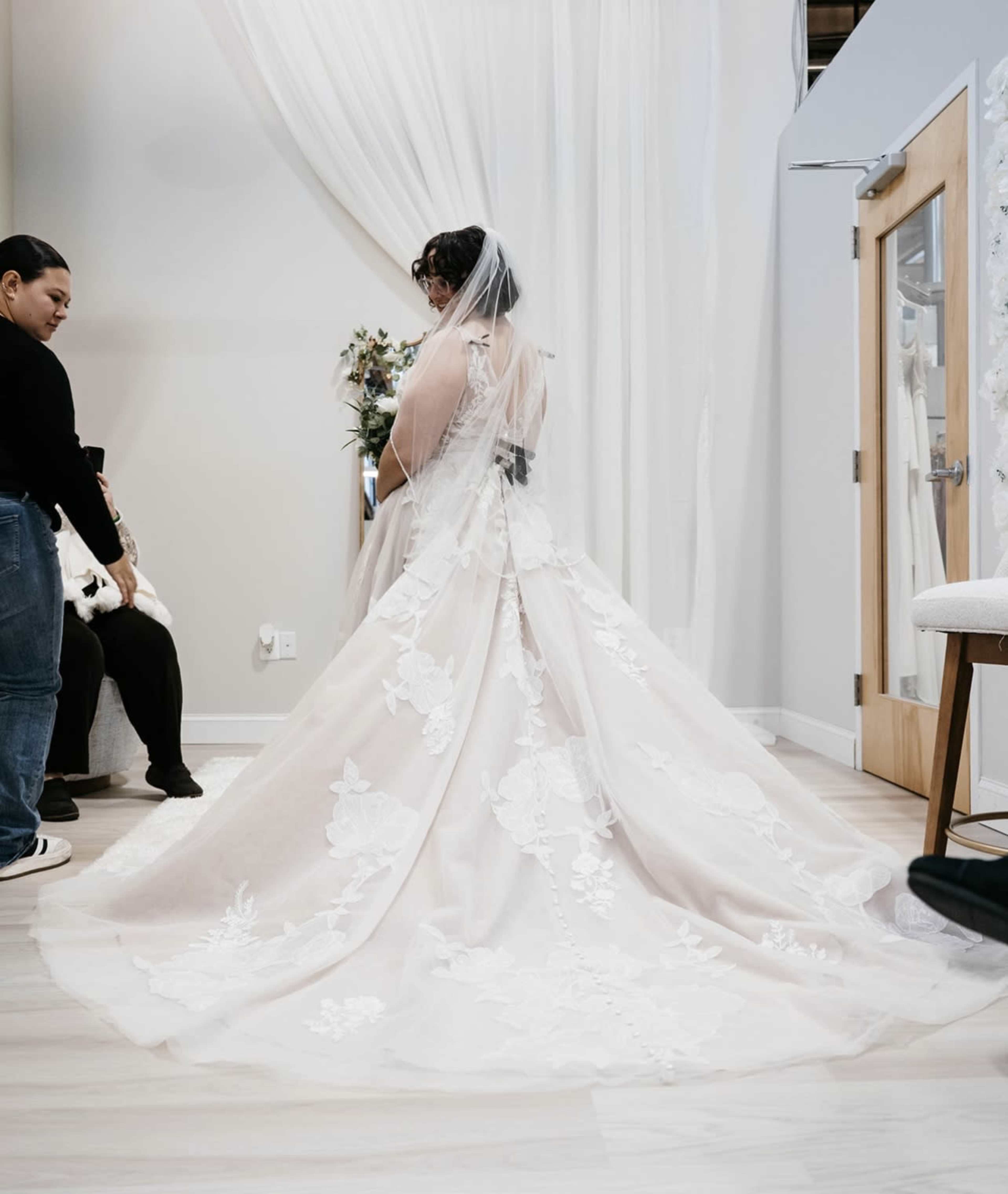 A bride in a wedding dress with floral appliqués stands with her back to the camera in a bridal shop, while two women sit nearby.