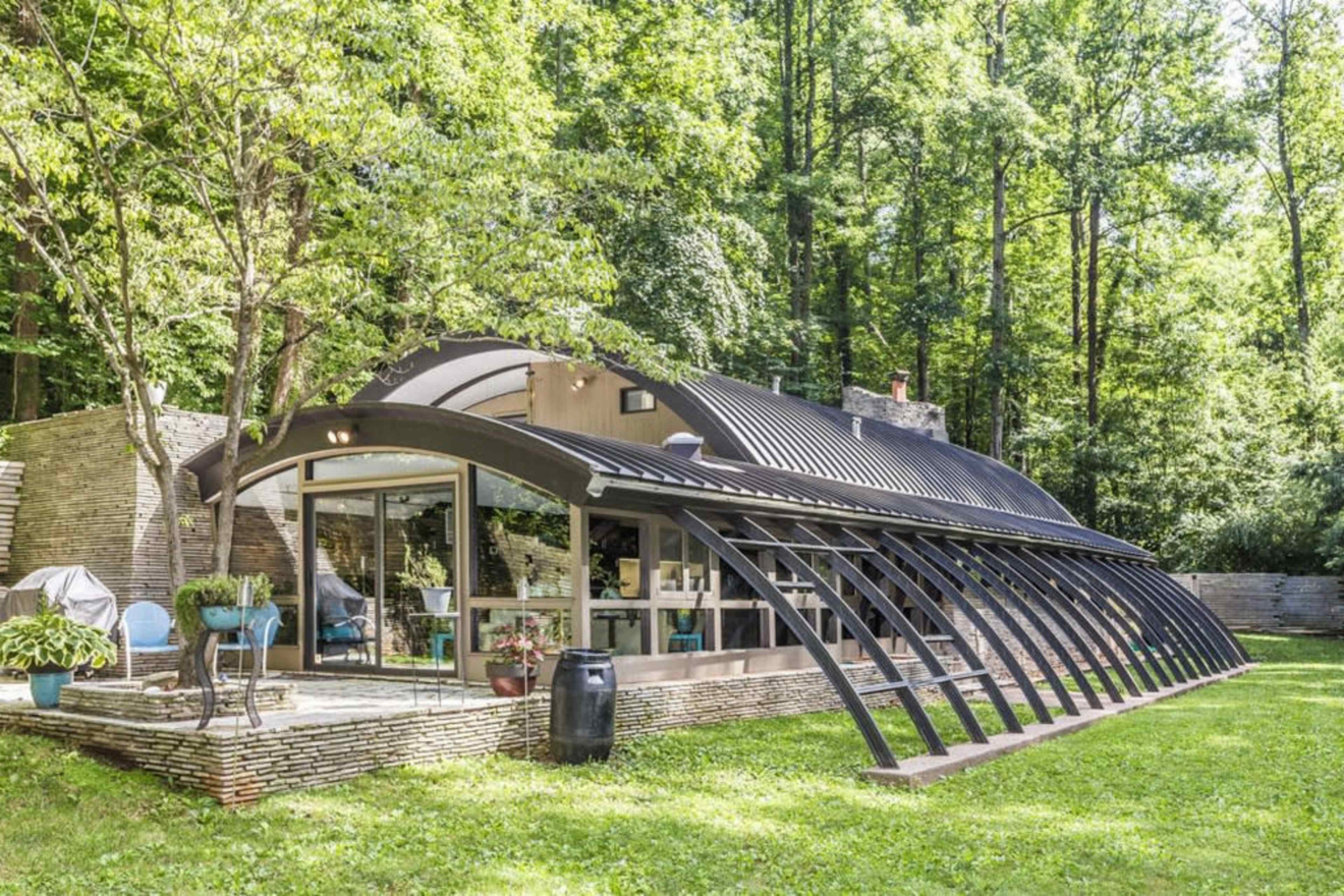 A modern, curved house with large glass windows surrounded by trees and a green lawn.
