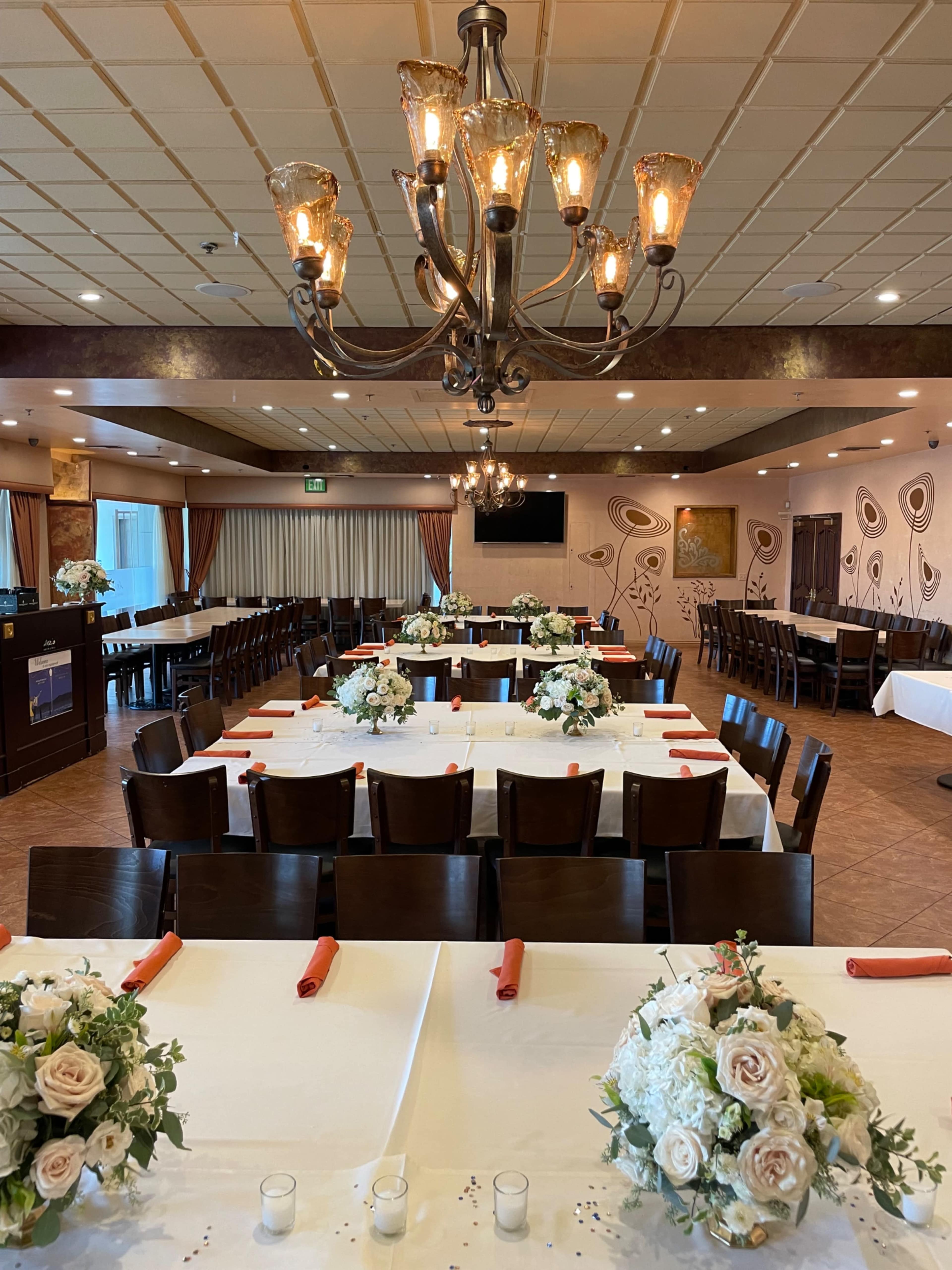 A dining room arranged with several tables set for a formal event, featuring floral centerpieces and a chandelier overhead.
