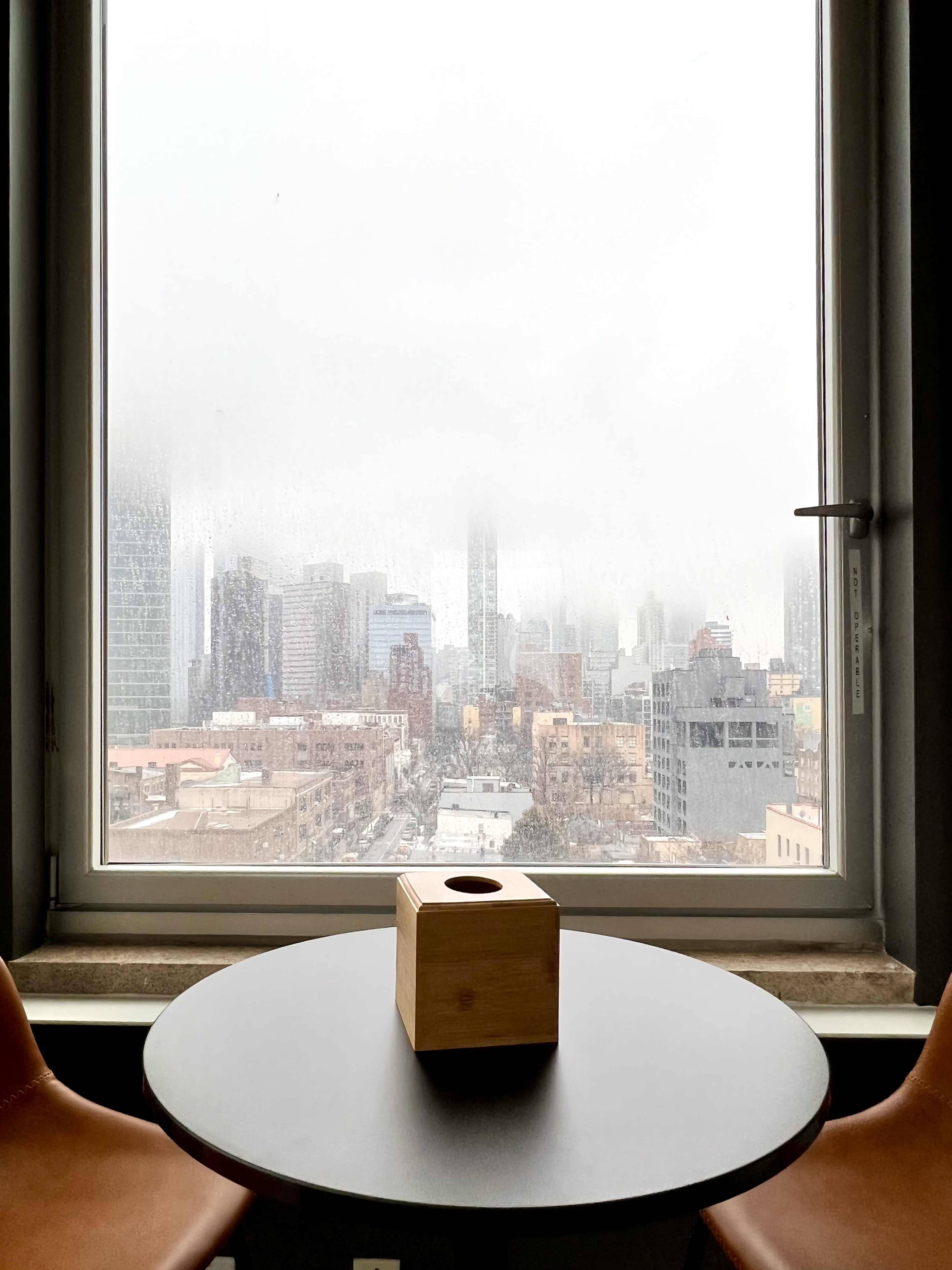 A wooden cube sits on a round table beside a foggy window overlooking a cityscape shrouded in mist.