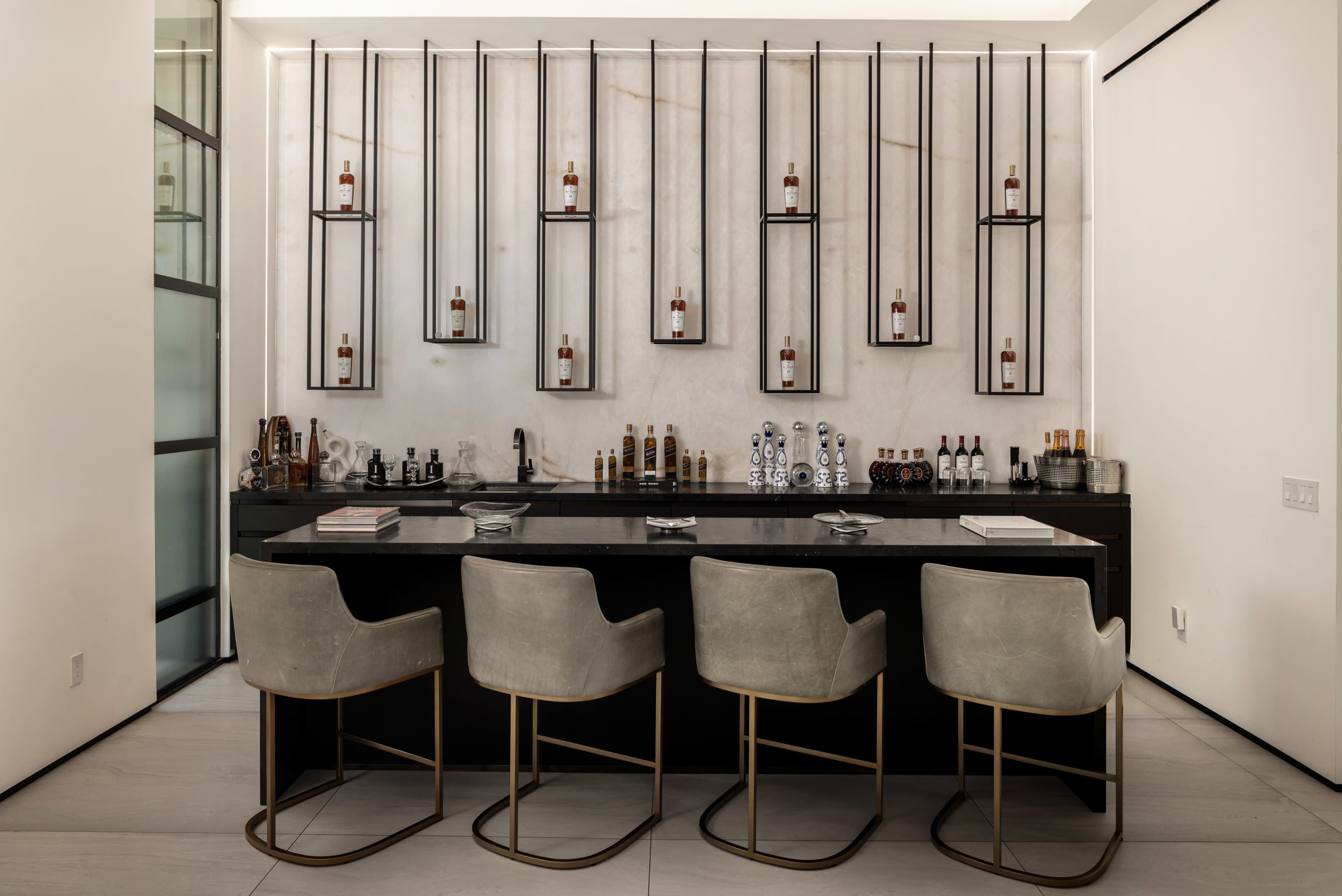 A sleek bar area features a black countertop, four upholstered stools, and a stylish display of various bottles arranged on wall-mounted shelves.