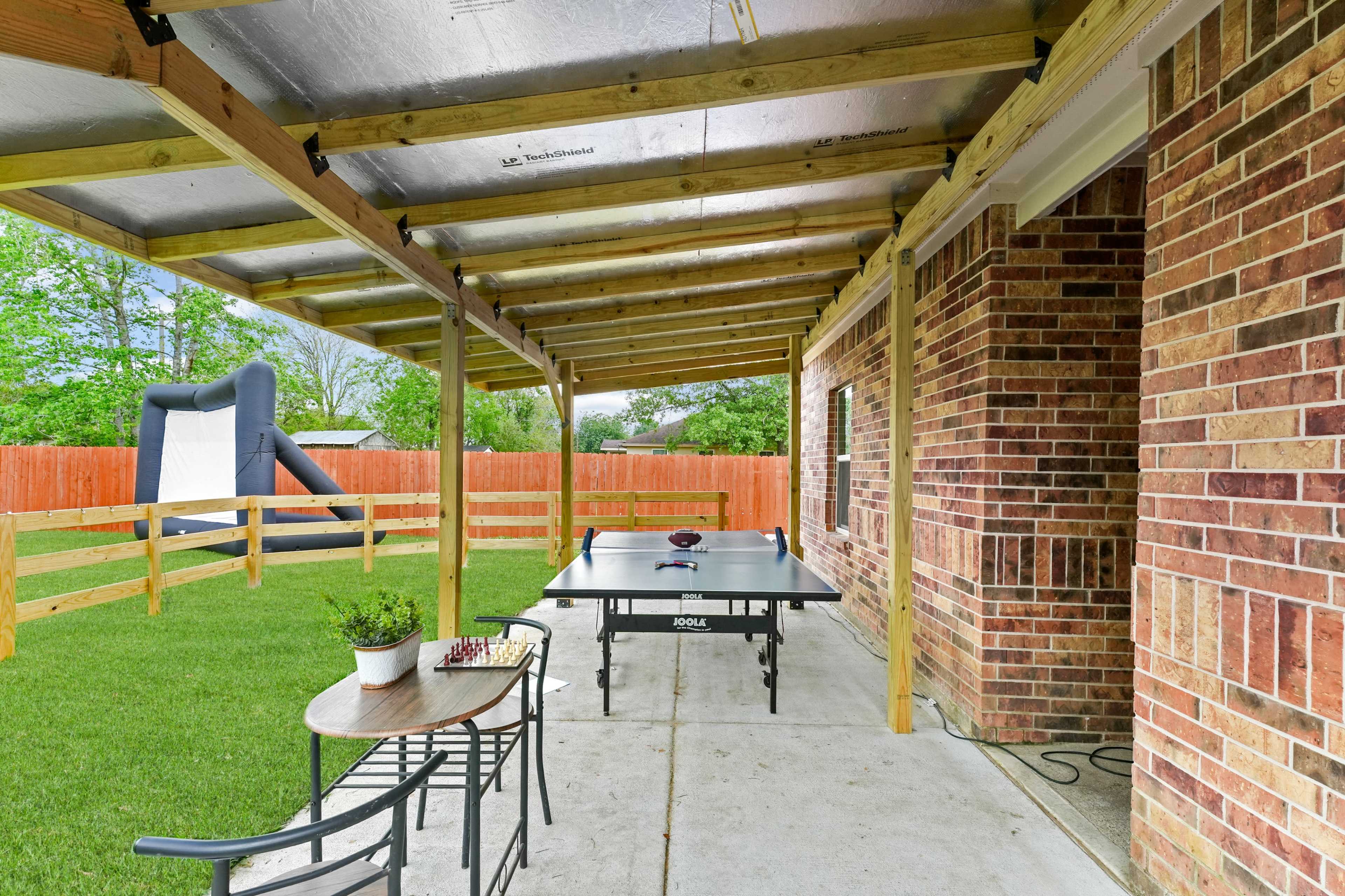 A covered patio area features a ping pong table and a small table with chairs, overlooking a grassy yard and a bounce house in the background.