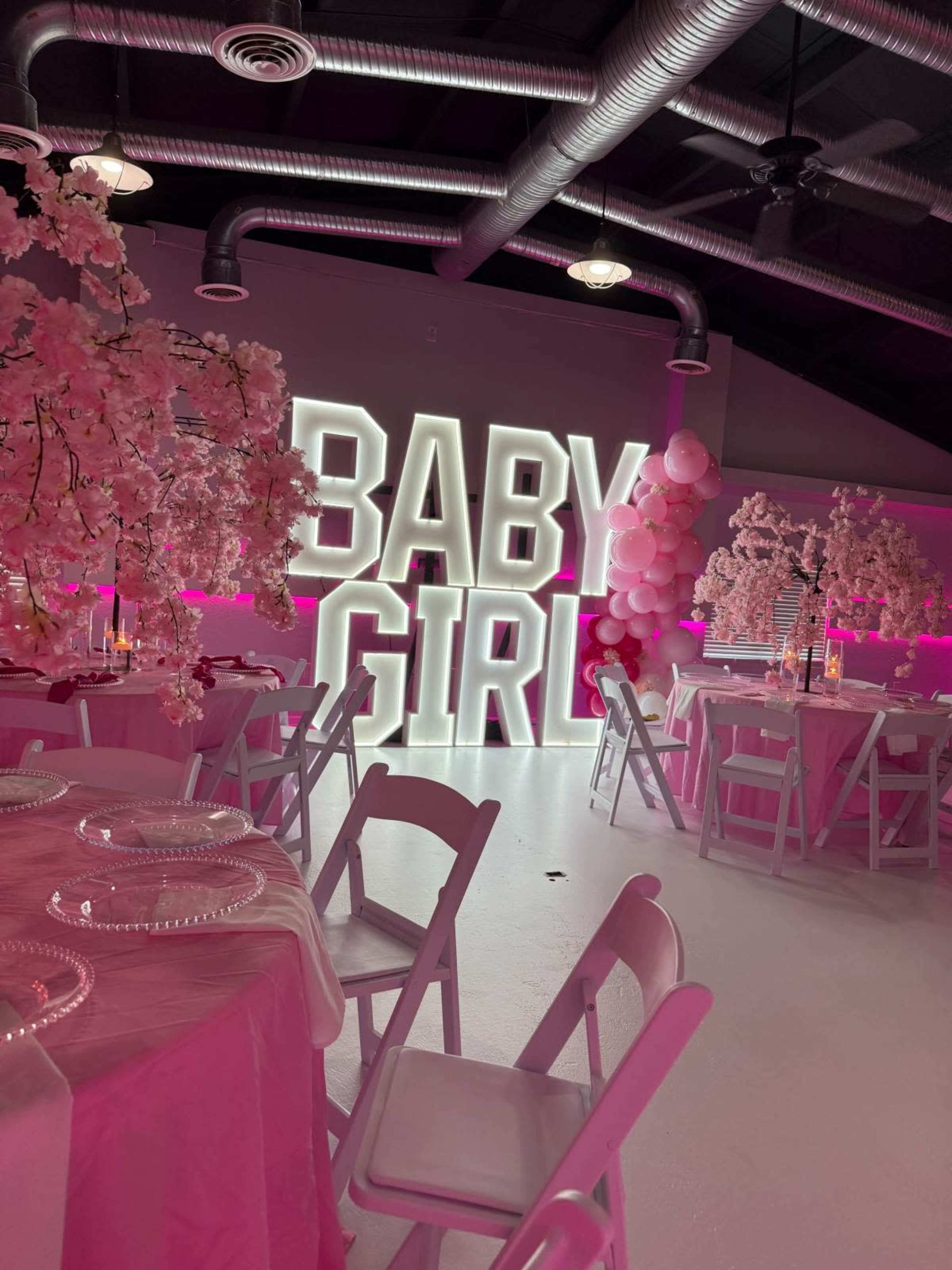 The image shows a celebratory event space decorated with pink balloons and artificial cherry blossom trees, featuring a large illuminated sign that reads "BABY GIRL."