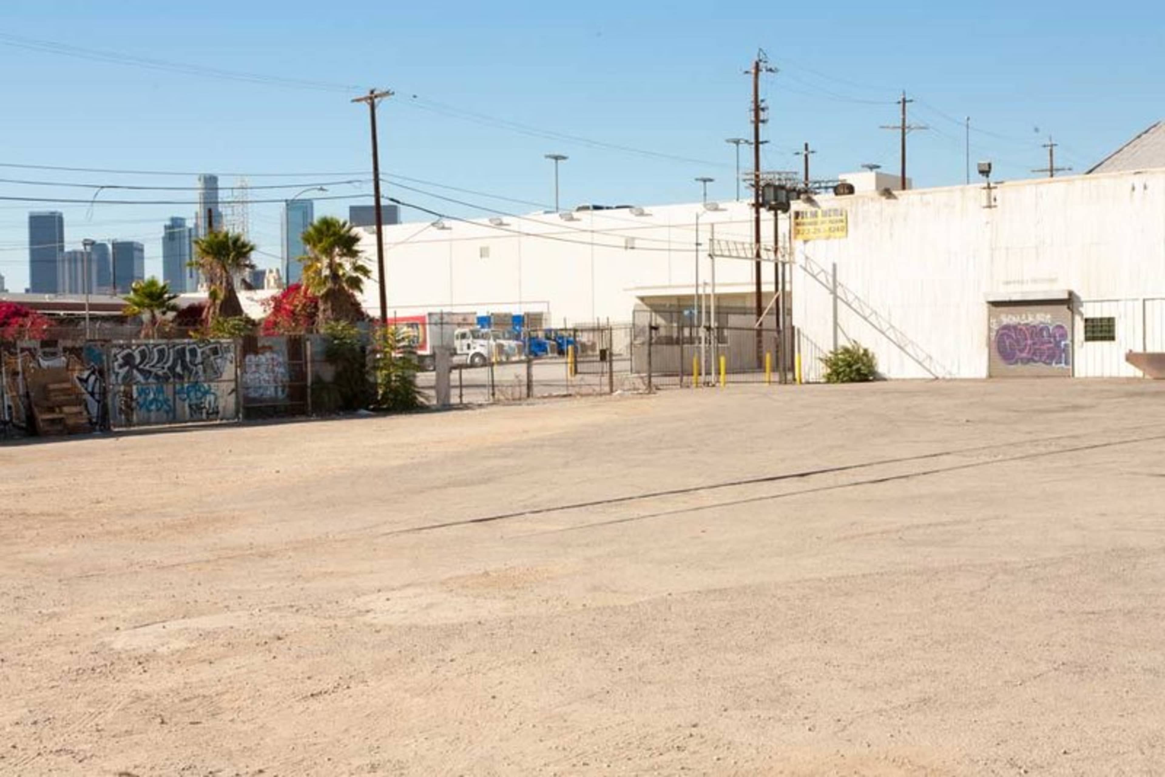 The image shows an empty dirt lot bordered by a chain-link fence, with a backdrop of industrial buildings and palm trees under a clear blue sky.