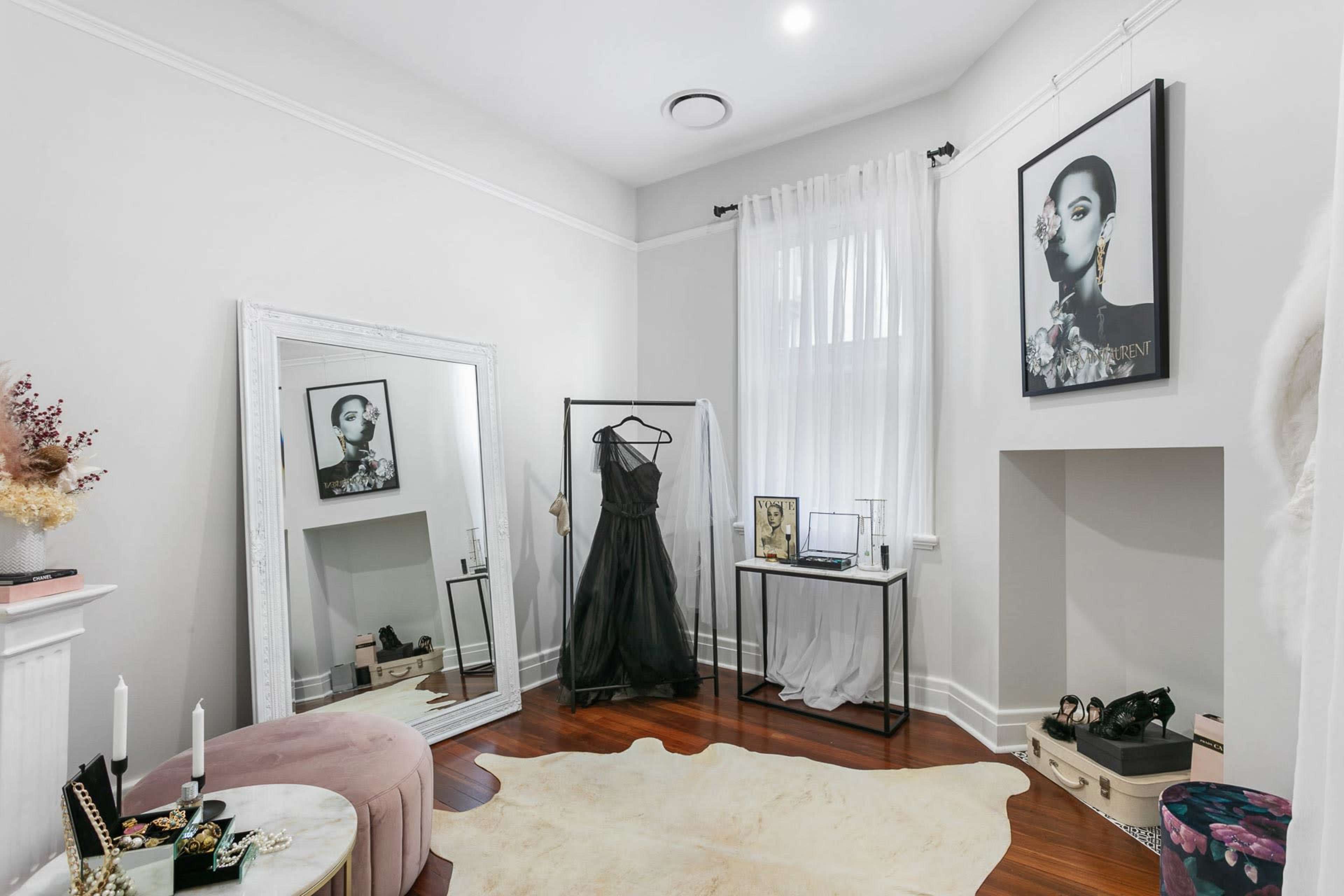 A corner of a stylish room featuring a mirror, a black dress on a clothing rack, and a decorative table with various items.