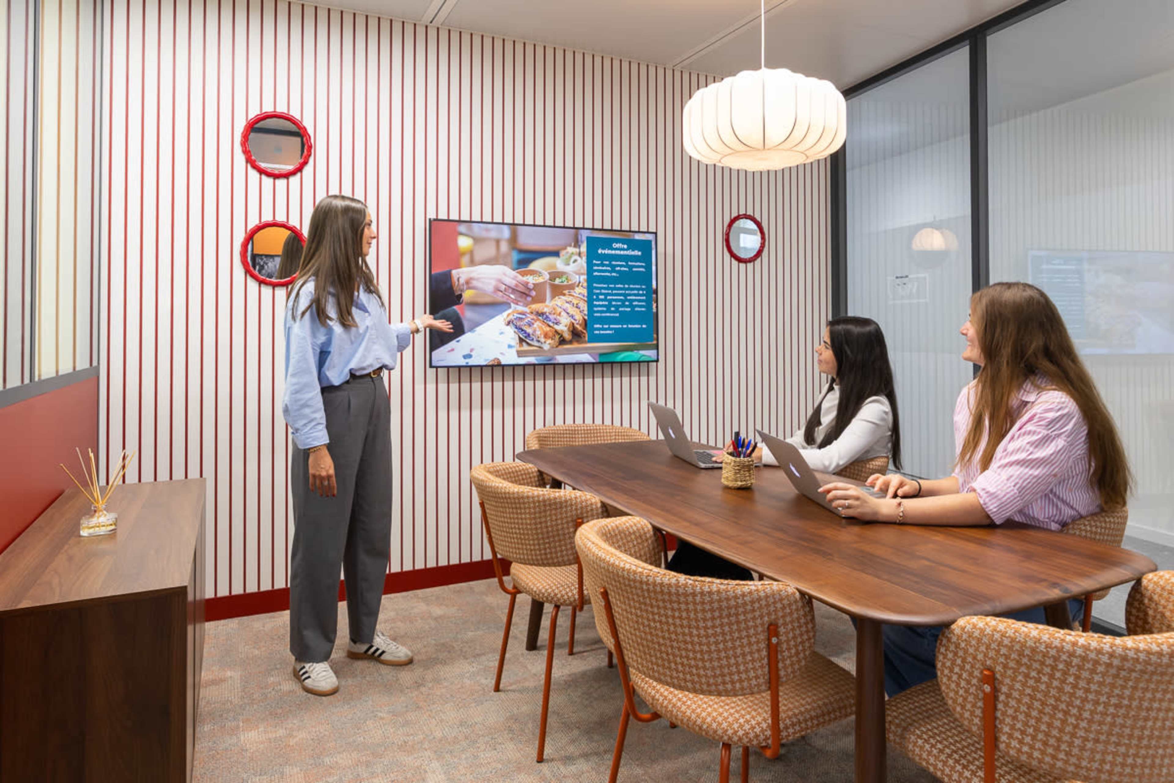 A woman is presenting to two seated colleagues in a modern conference room with a large screen displaying information.