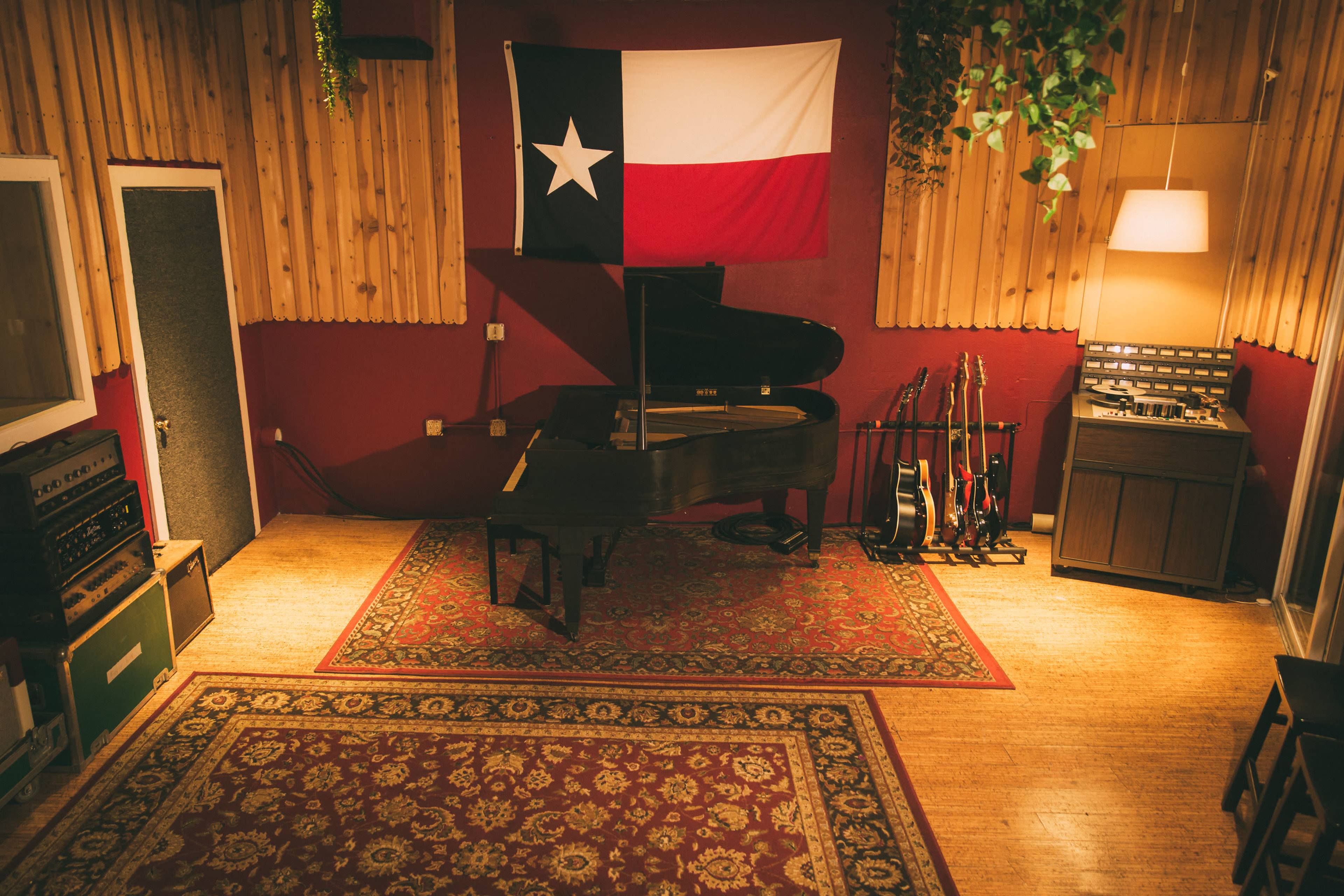A music studio interior featuring a grand piano, a Texas flag, various guitars, and recording equipment against wooden paneling.