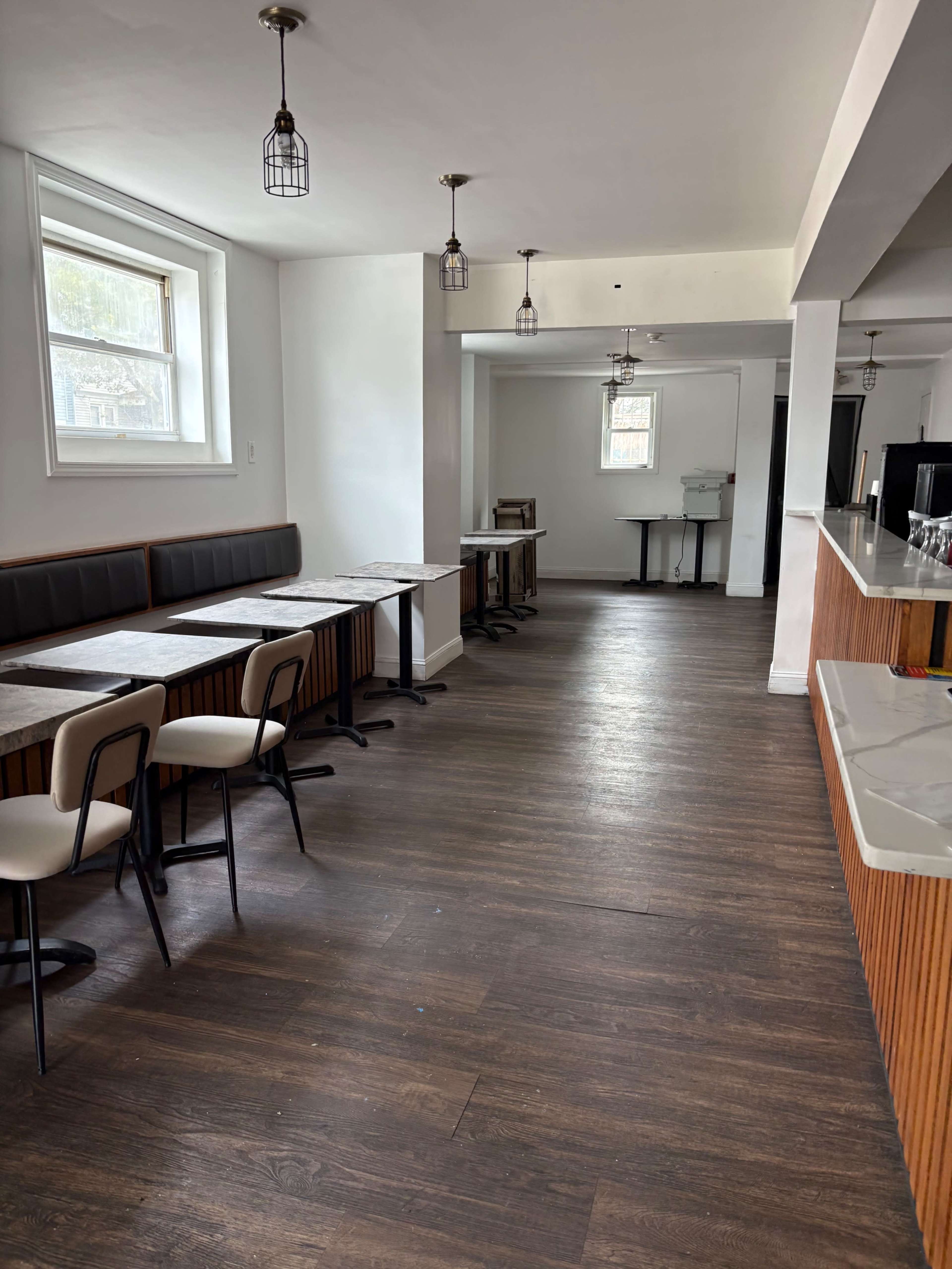 The image shows a sparsely furnished dining area with several tables and chairs along one side and a bar counter on the opposite side.