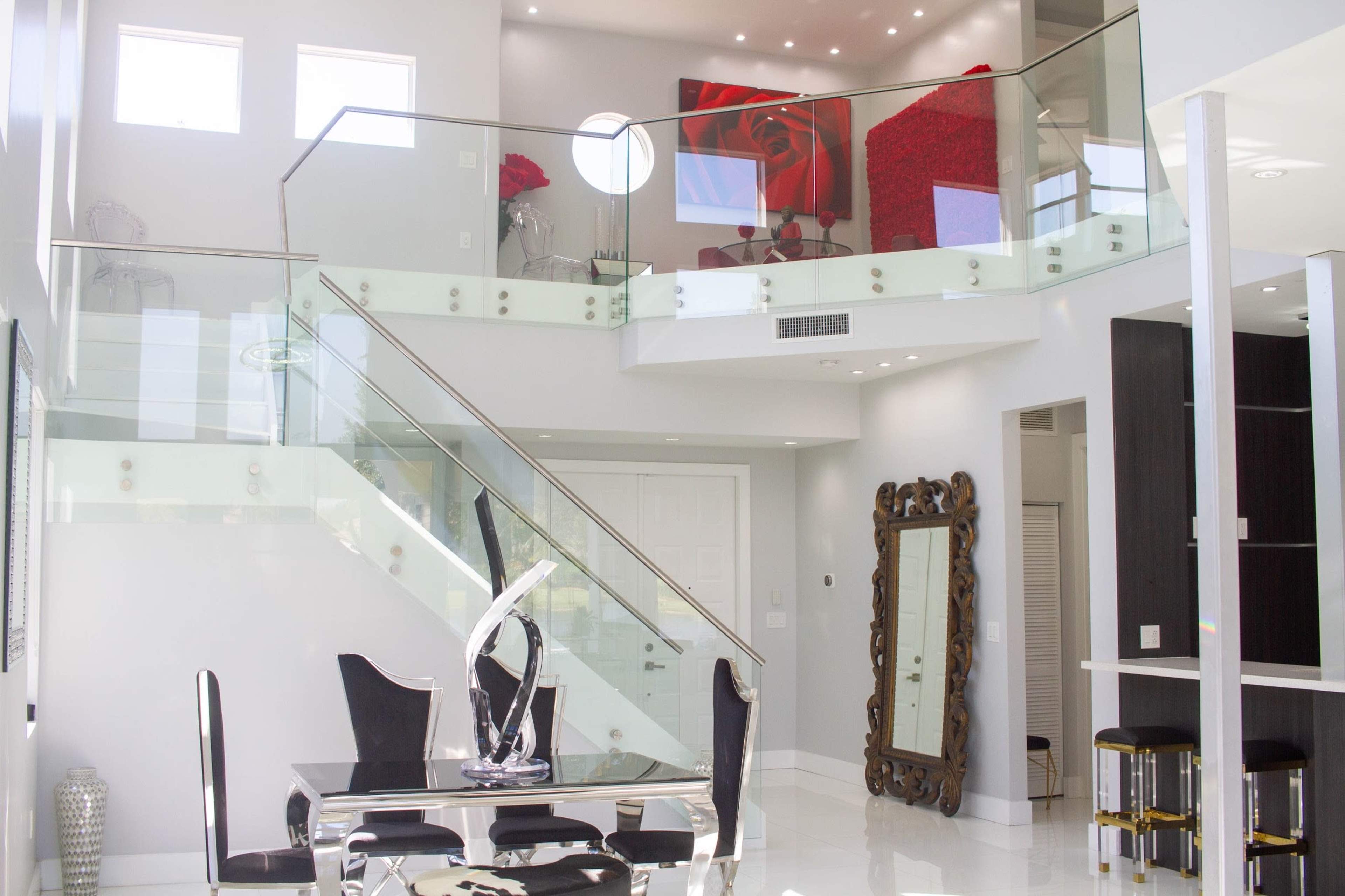 A modern, two-story living space features a glass staircase, sleek dining furniture, and a large abstract painting on the wall.
